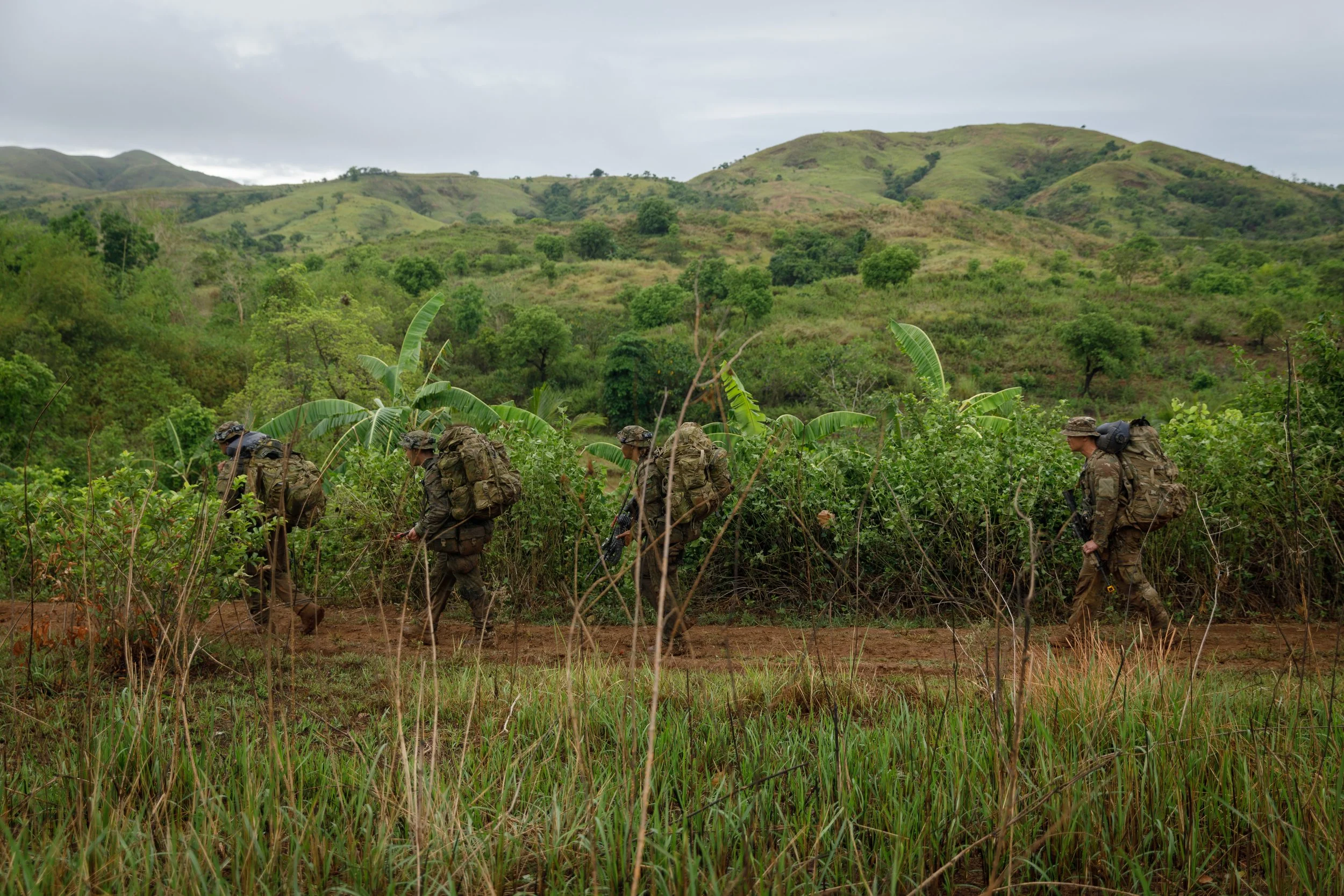 Four soldiers in camouflage uniforms and backpacks walking through a green, hilly, tropical landscape with banana plants and tall grass.