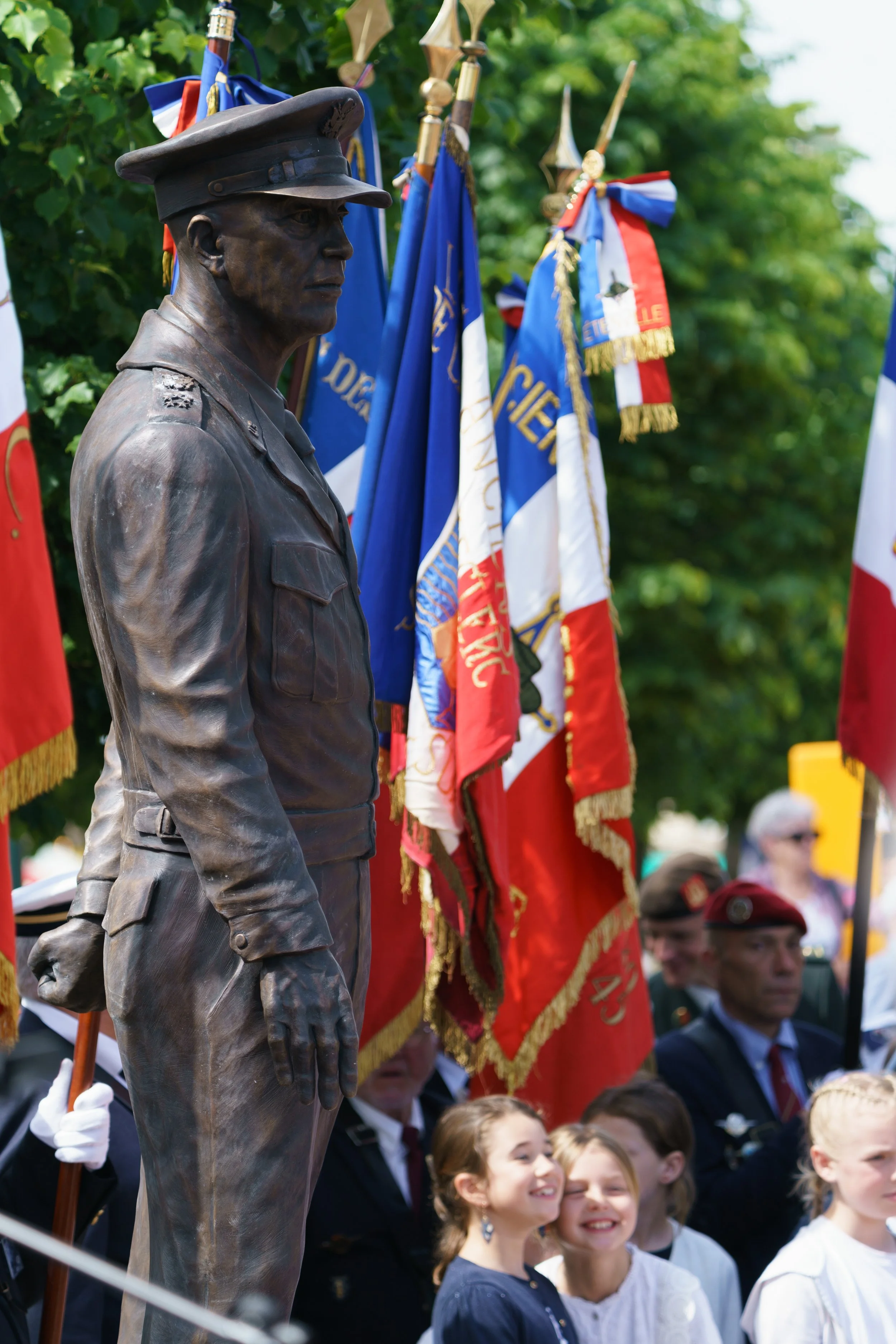 A bronze statue of a man in a military uniform with a cap, set in front of a display of colorful Franco-American flags during a parade or ceremony, with children and adults in the background.