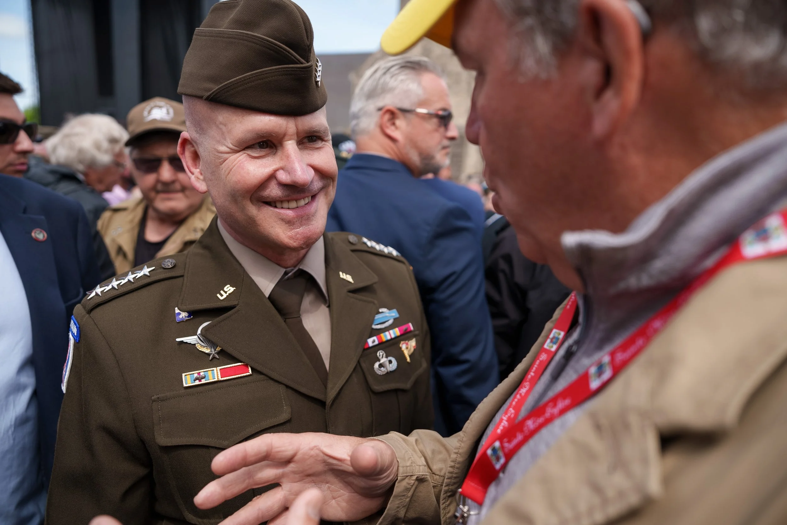 A smiling U.S. military officer in uniform engaged in a handshake and conversation with another person at a public event, with several onlookers in the background.