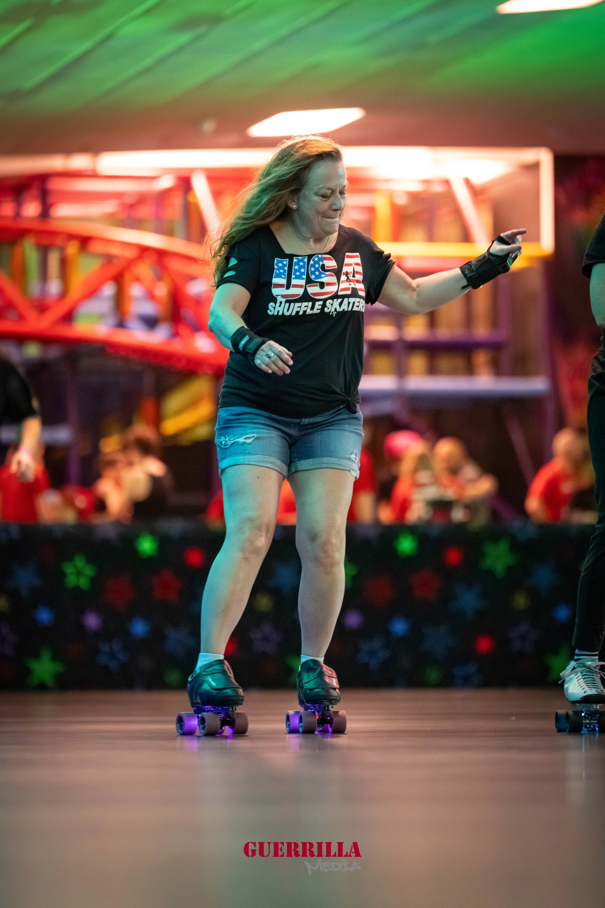 A woman roller skating indoors under colorful lighting. She is wearing a black USA Shuffle Skater t-shirt, denim shorts, and protective wrist guards. The background shows a vibrant, lively atmosphere with people and colorful decorations.