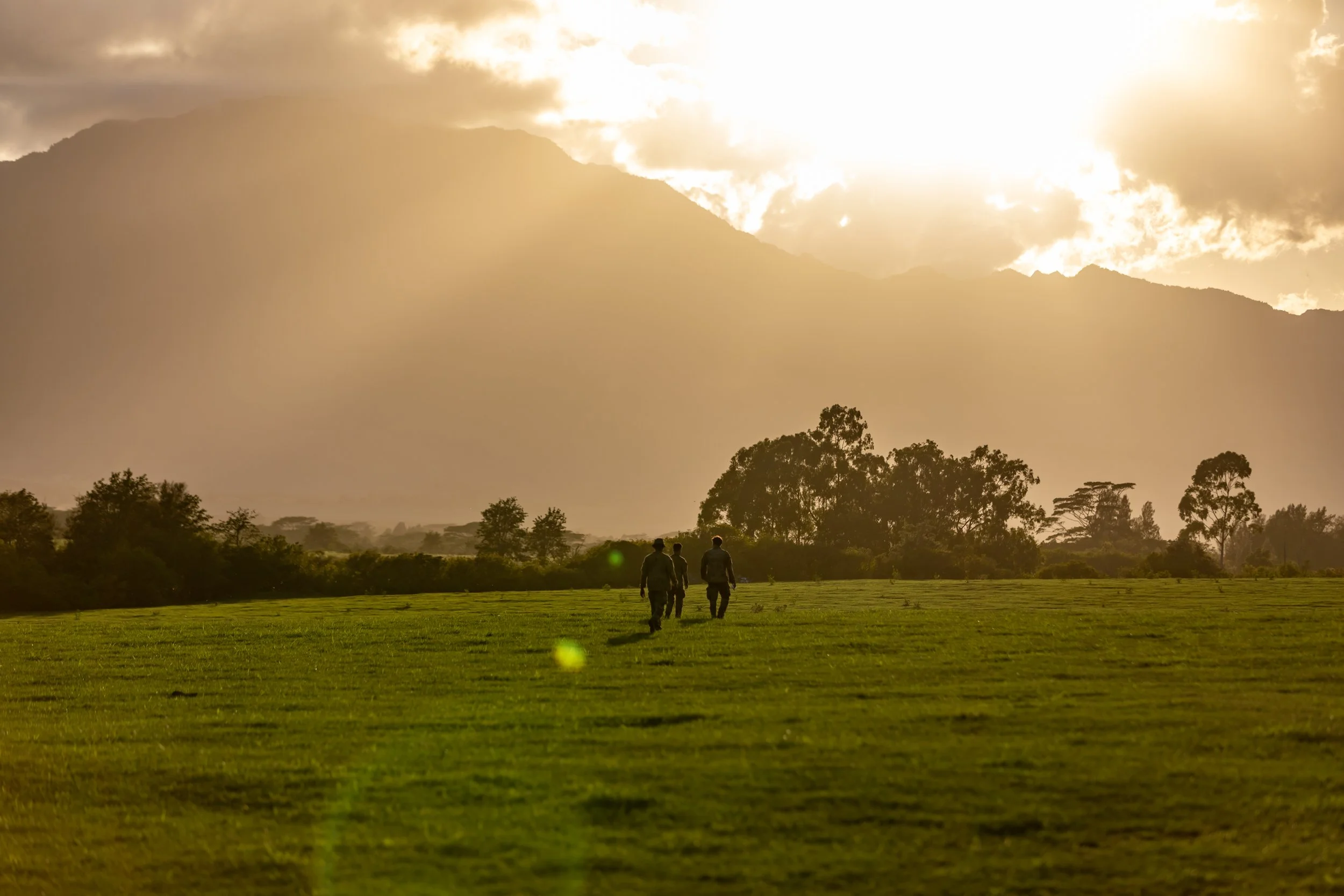 Three soldiers walking across a grassy field toward a distant mountain range with partly cloudy sky during sunset.