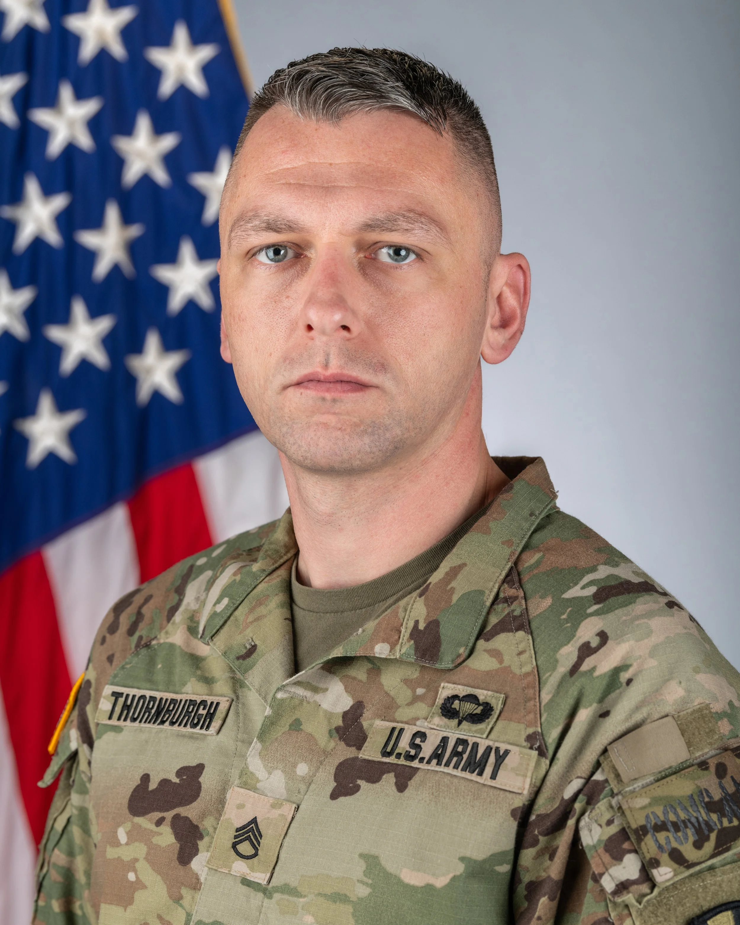 A U.S. Army soldier in camouflage uniform standing in front of an American flag.