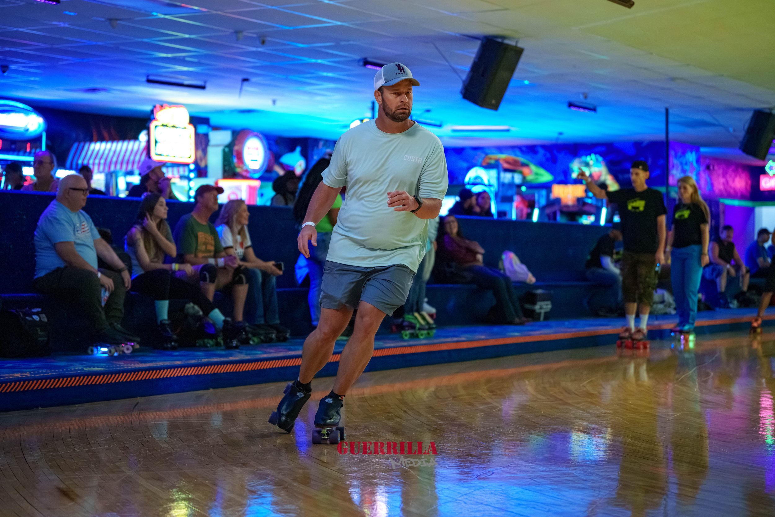 A man roller skating on an indoor rink with neon and LED lights, with people sitting on benches along the wall and others skating in the background.
