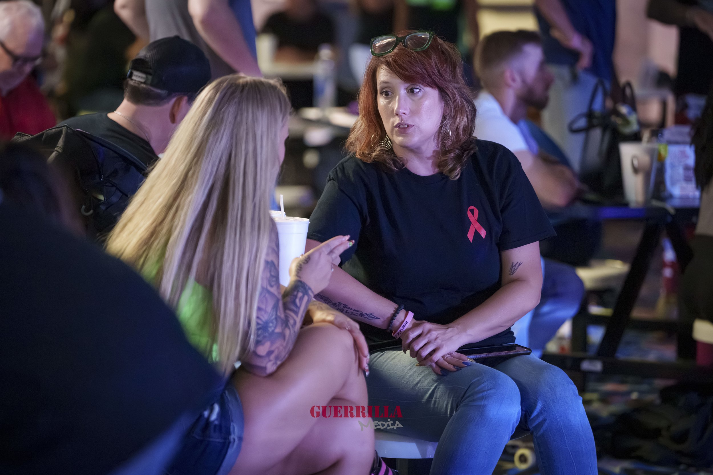 A woman with red hair, wearing a black t-shirt with a pink ribbon, is talking to a blonde woman at a social event. The woman with red hair has glasses on her head and tattoos on her arm.