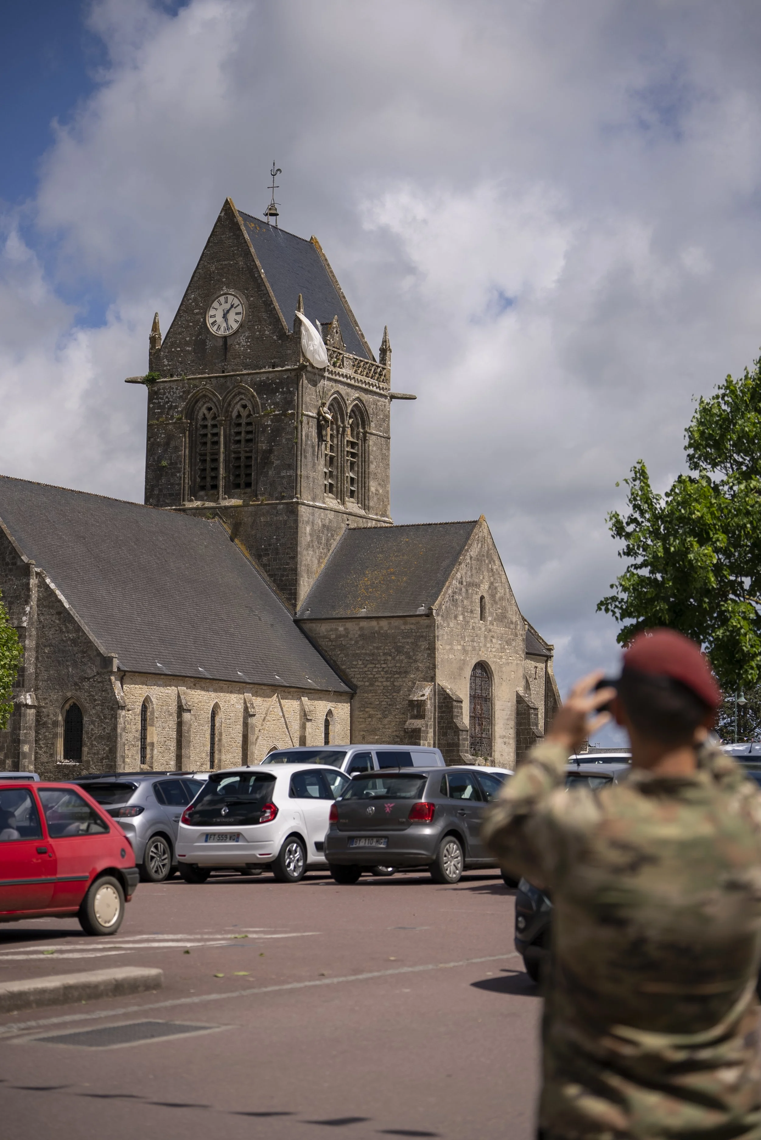 A church with a dark stone exterior and a tall clock tower, parked cars in front, and a soldier in camouflage uniform with a red beret standing and saluting or shielding eyes, with a partly cloudy sky overhead.