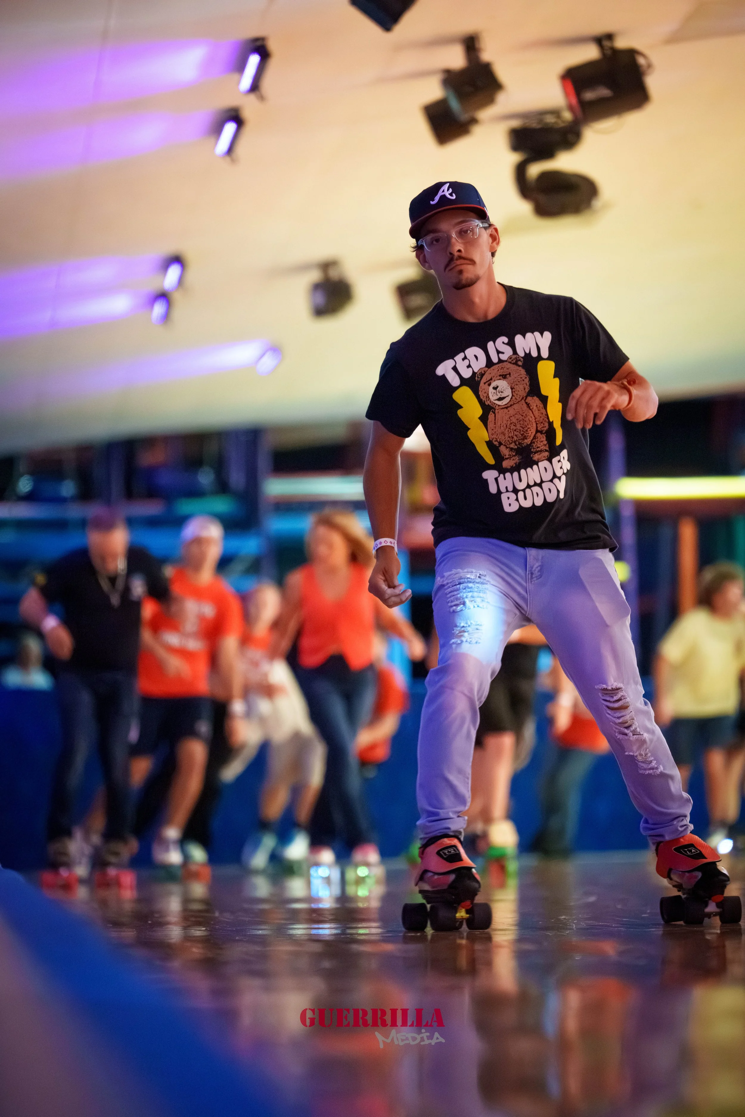 A man roller skating indoors at a skating rink with colorful lighting. He is wearing a black T-shirt that says 'TEDIS IS MY THUNDER BUDDY' with a bear graphic, ripped jeans, a baseball cap, and glasses. There are people in the background also skating