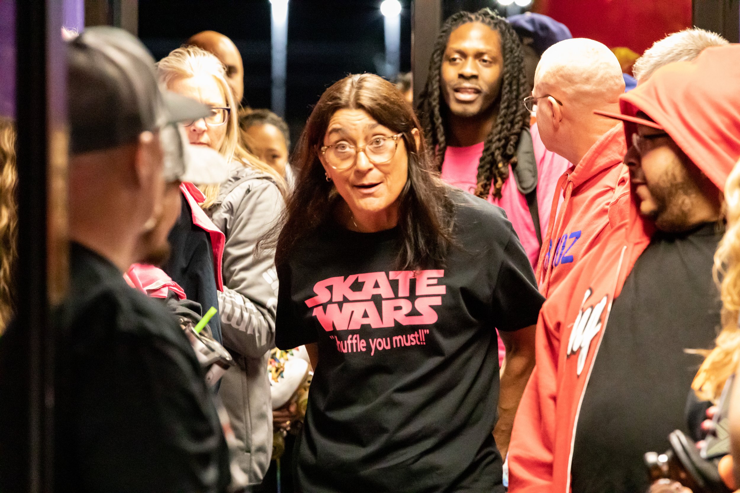 A woman wearing a black shirt that says "Skate Wars" is talking to a group of people at a gathering inside a building. People around her are listening and wearing casual jackets and hoodies.