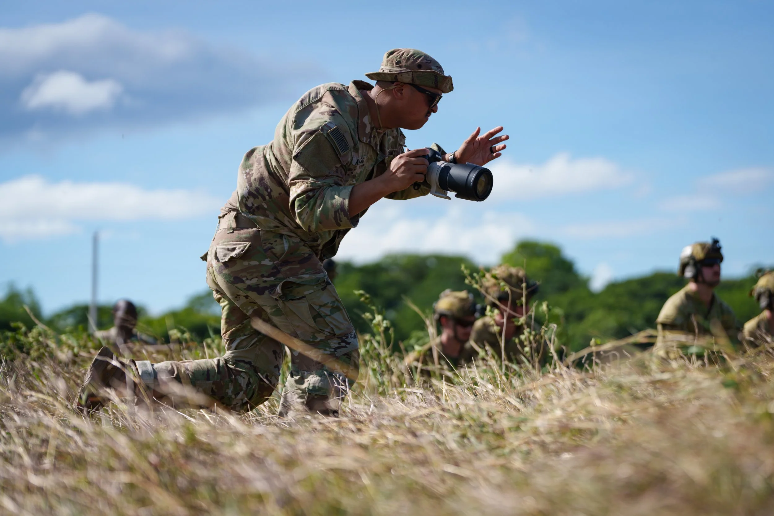 Soldiers in camouflage uniforms crawling on the ground during a training exercise, with one holding a camera or spotting scope, under a blue sky with some clouds.