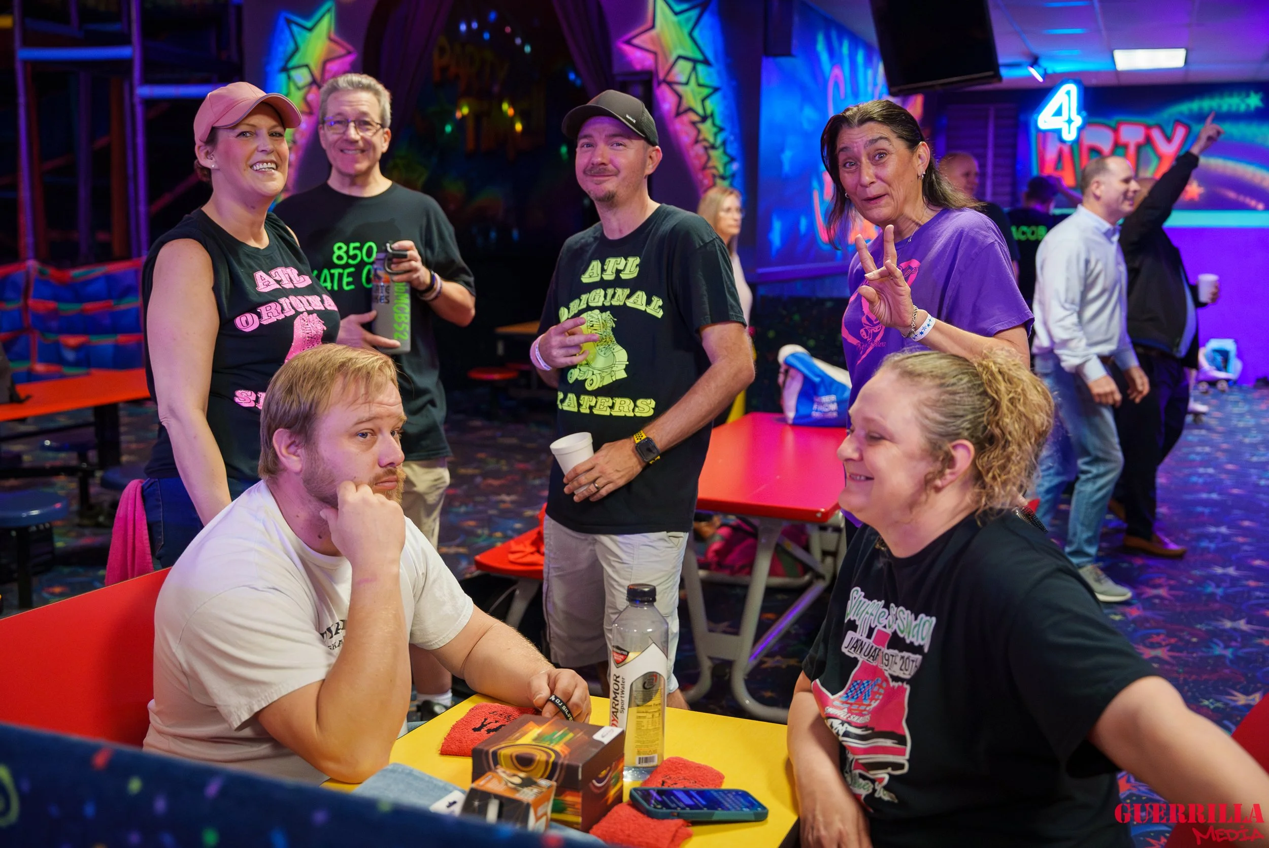 Group of people at an indoor bowling alley with colorful neon and LED lights, some sitting at a table, others standing and posing for the photo, with a sign that says '4 Party' in the background.
