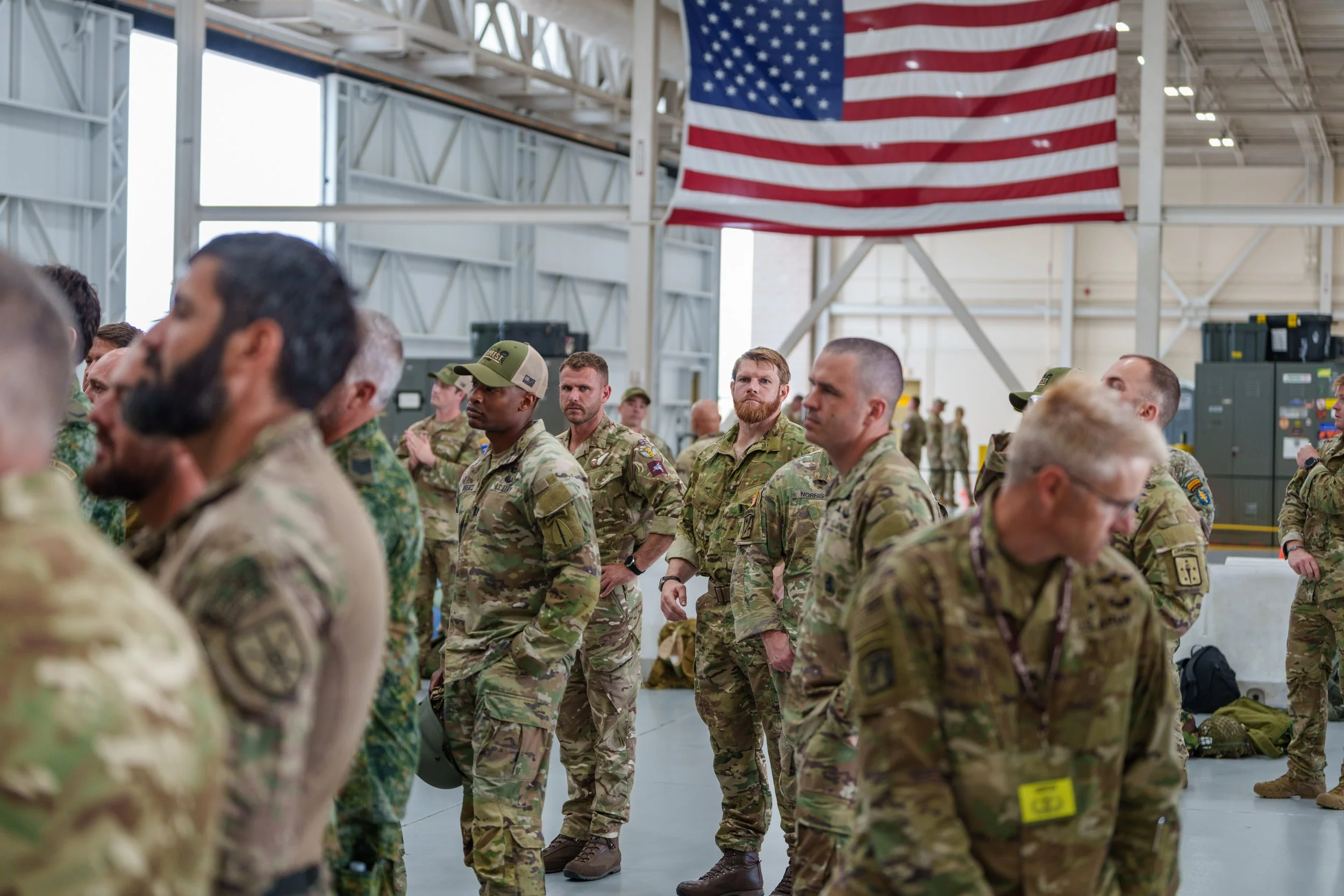 Military personnel standing in formation inside a hangar with an American flag hanging from the ceiling.