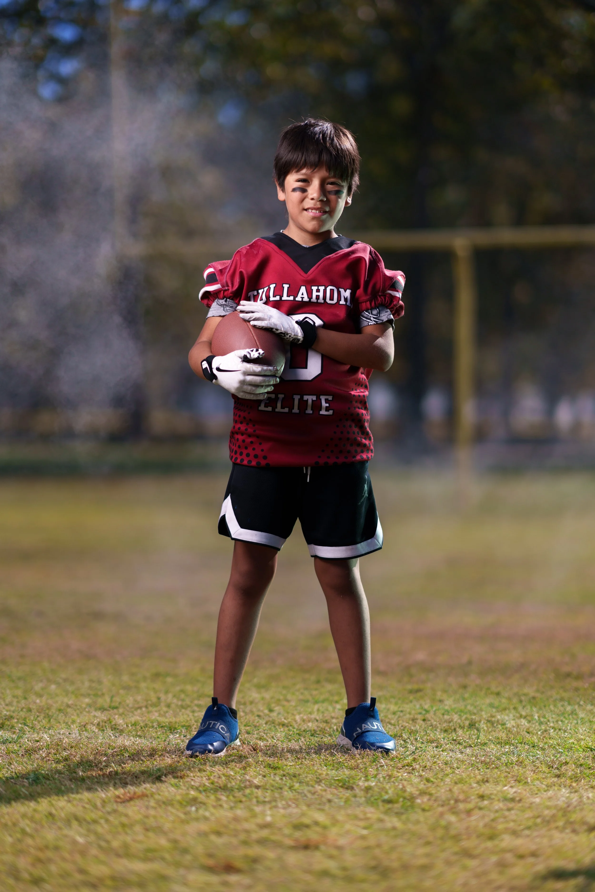 Young boy in football uniform holding a football on a grassy field with trees in the background.