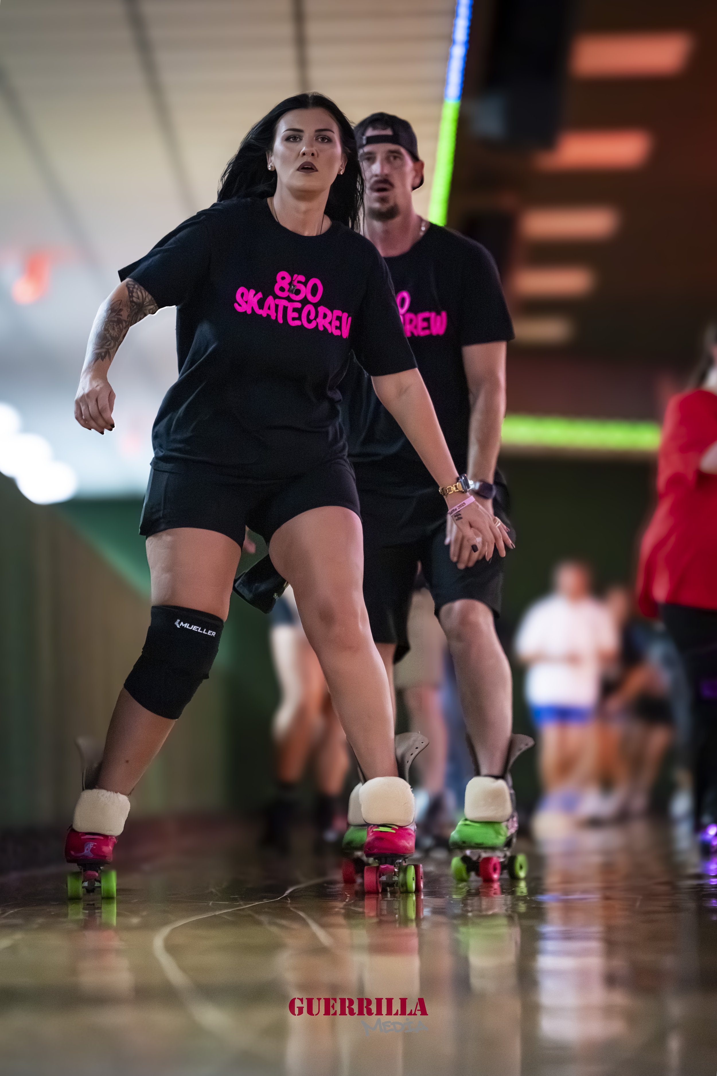 Two people roller skating indoors, wearing black shirts with pink text, with a woman in the front and a man behind her. The woman has long black hair and a tattoo on her left arm, and the man has a beard and is wearing a cap backwards.