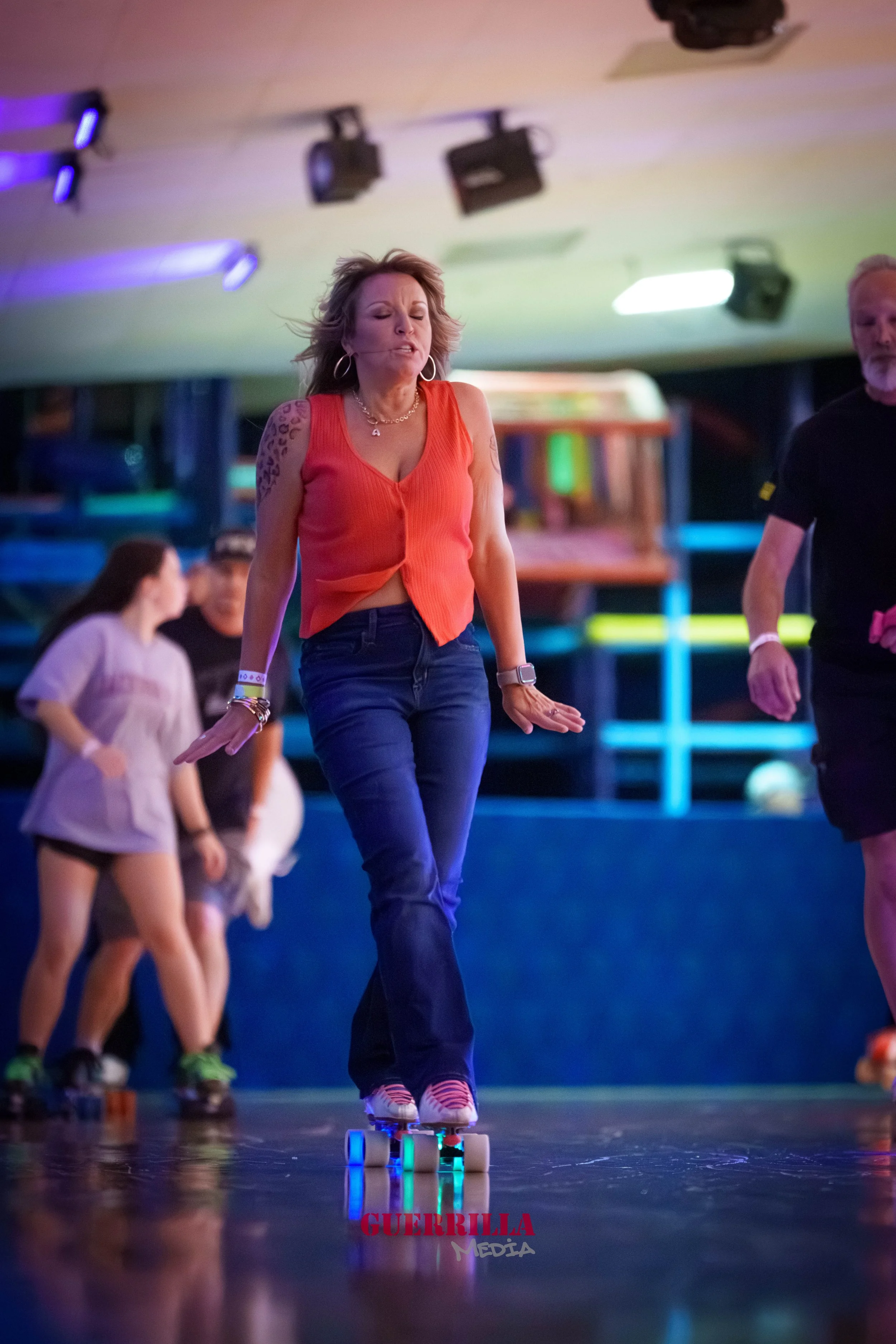 A woman roller skating in an indoor skating rink, wearing a sleeveless orange top and dark jeans, with a serious expression on her face, surrounded by other skaters.