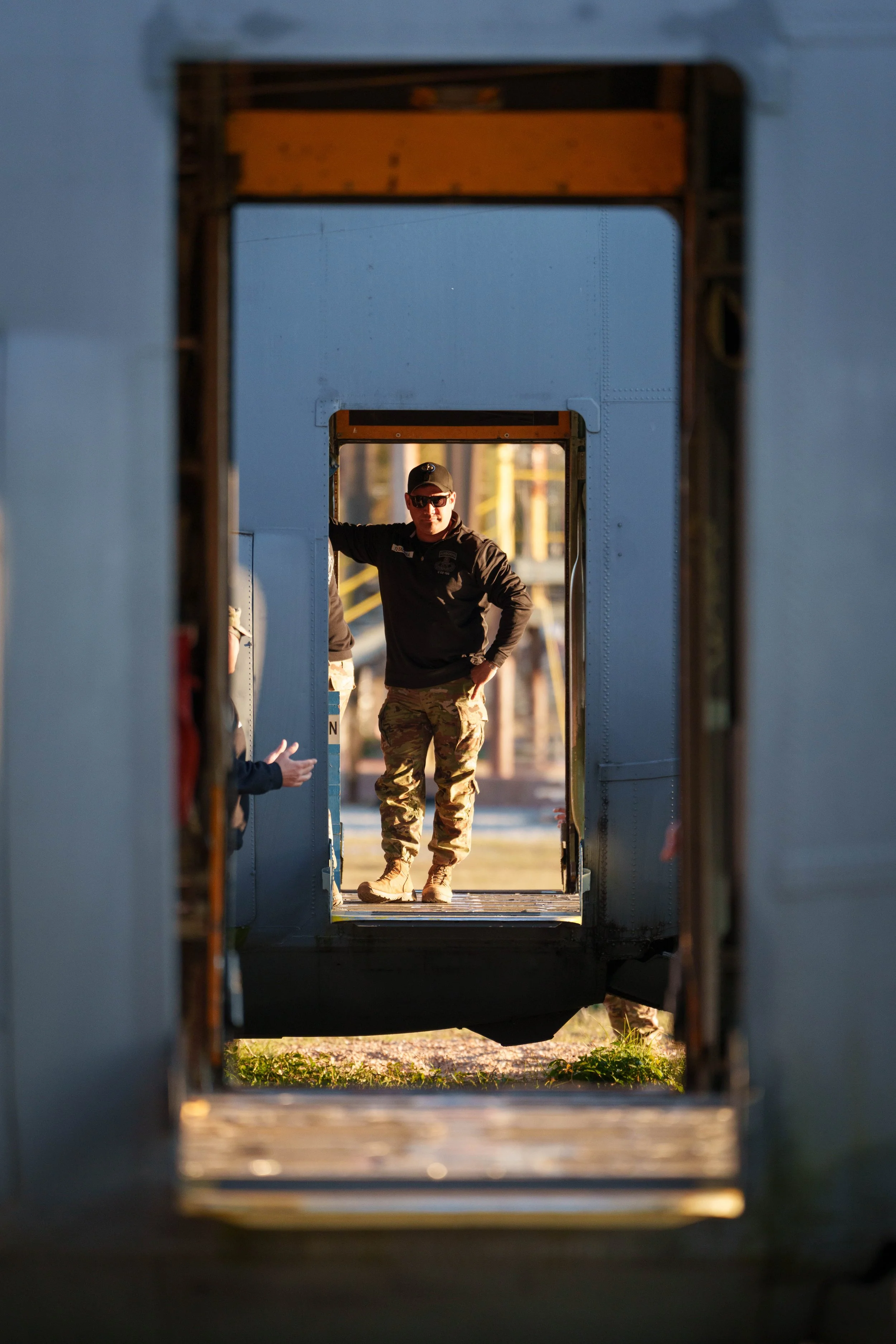 A military personnel, dressed in camouflage pants and black jacket, stands inside a blue aircraft doorway, seen through a series of door frames during sunset.
