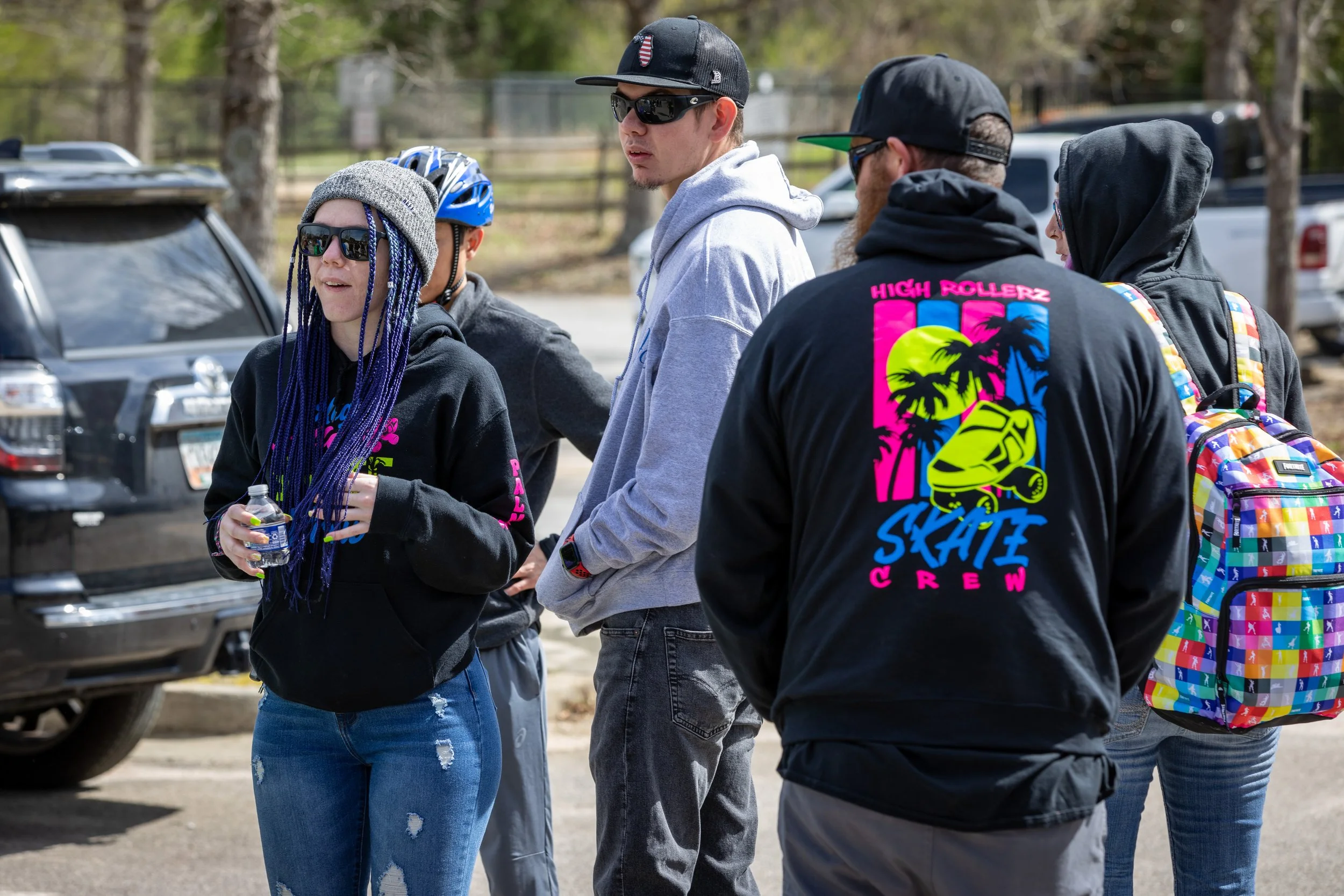 Group of young people standing outdoors, some wearing skateboarding apparel and accessories, in a parking lot with trees and vehicles in the background.