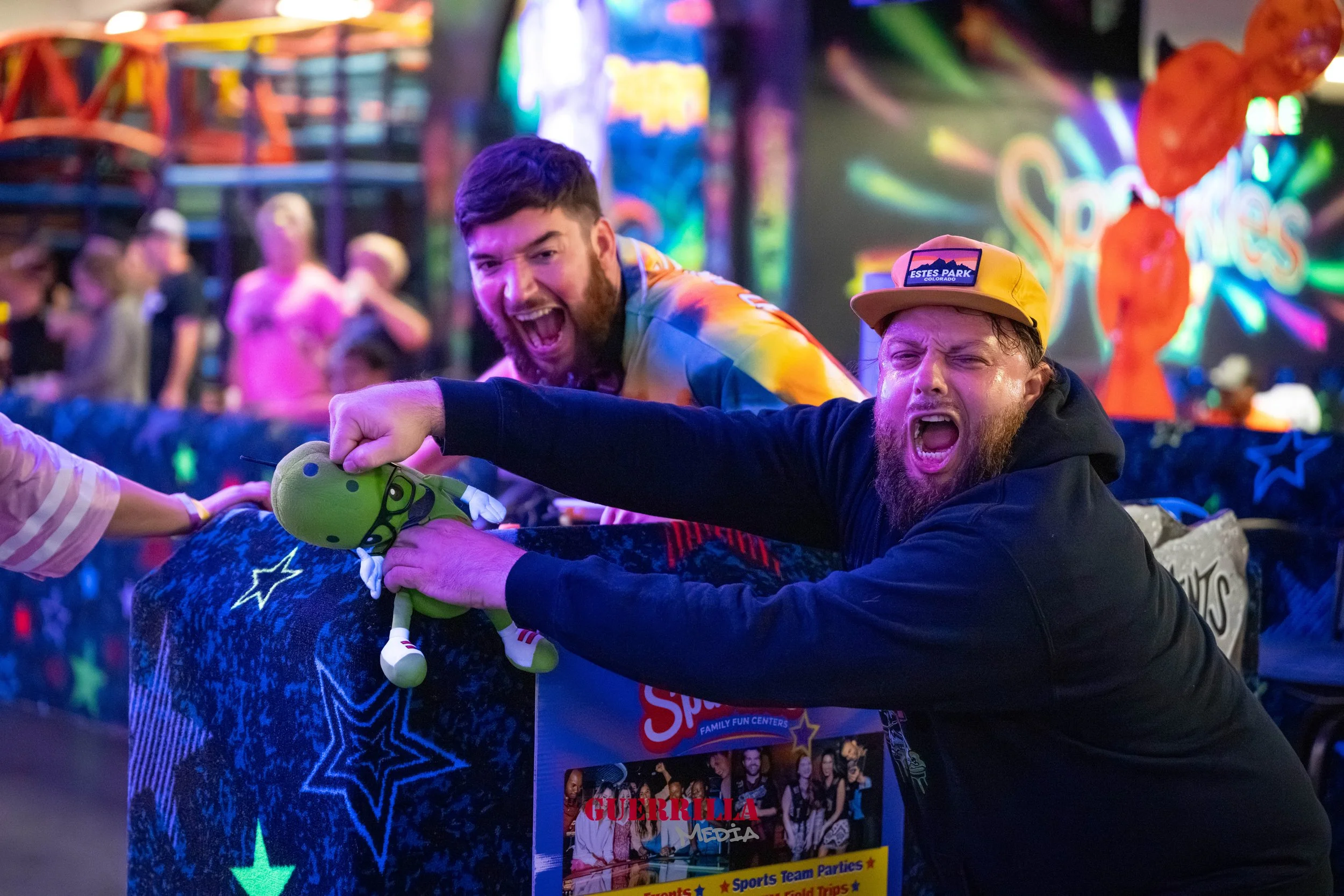 Two excited men playfully interact at an indoor arcade or fun center, one holding a plush toy with a green dinosaur or dragon design, both smiling and making expressive faces. Colorful neon lights and decorations illuminate the background.
