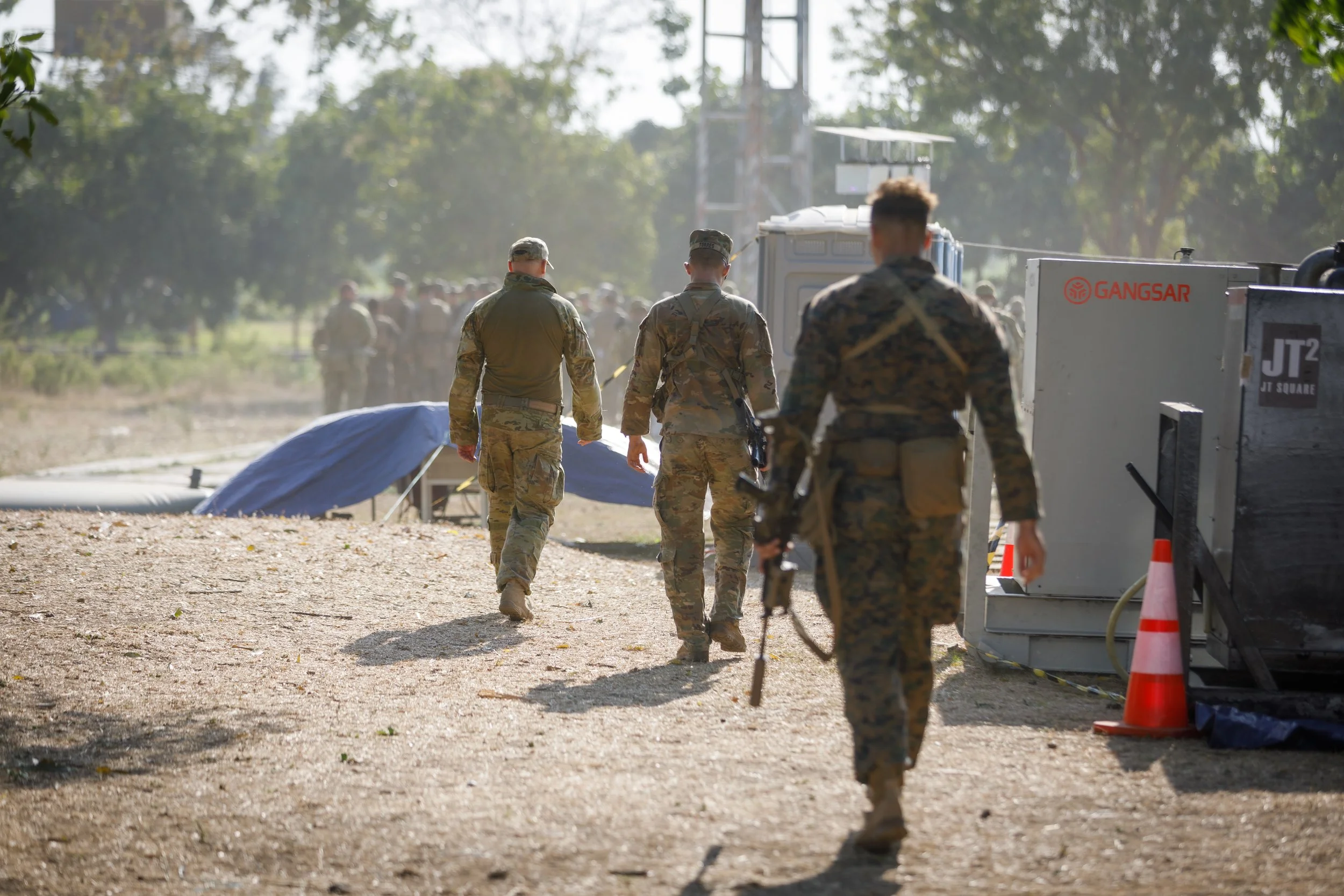 Group of soldiers walking away from the camera in a military setting, with equipment and trees in the background.