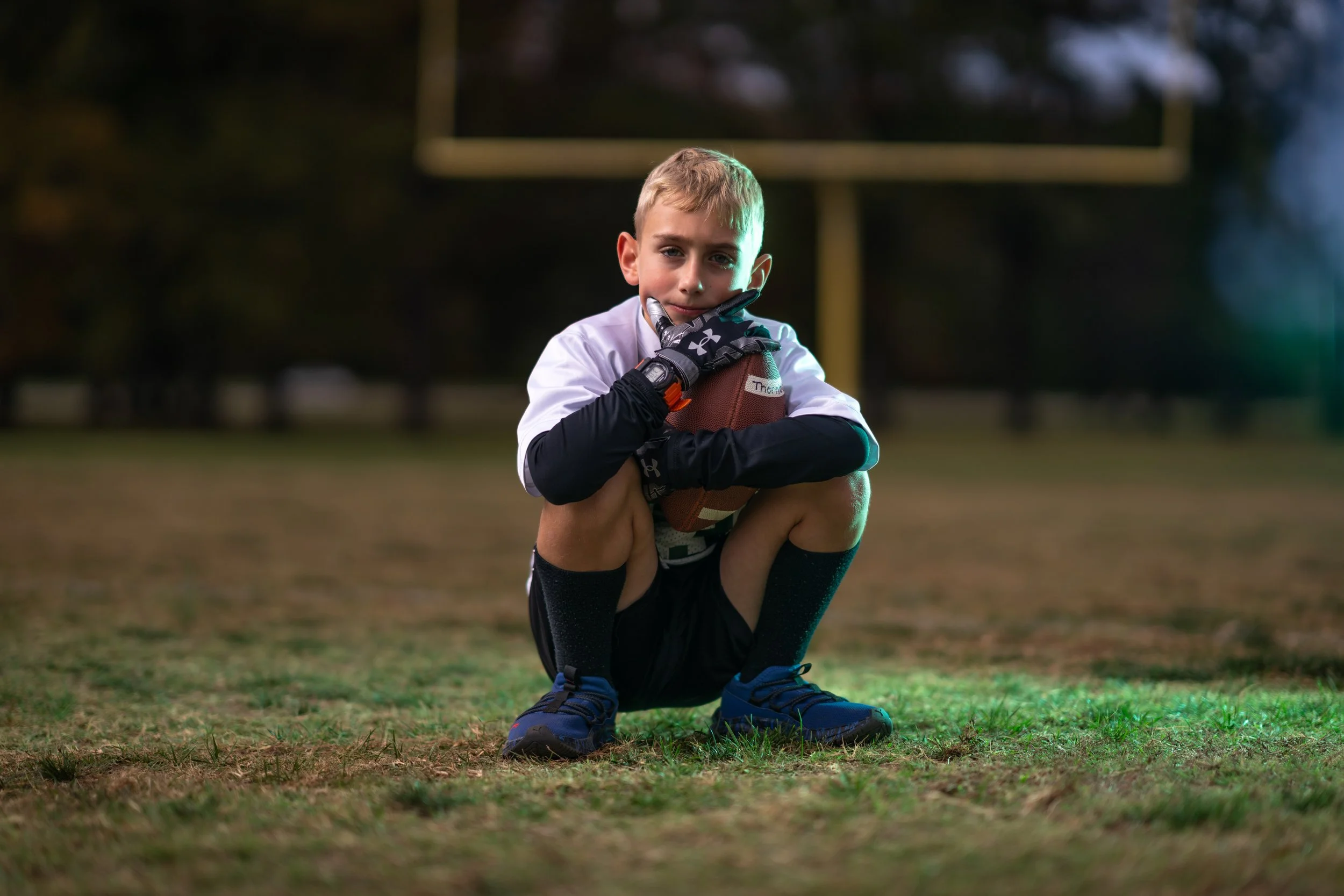 Young boy kneeling on a football field, holding a football, wearing football gloves, black shorts, blue shoes, and long black socks, with a football goalpost in the background.