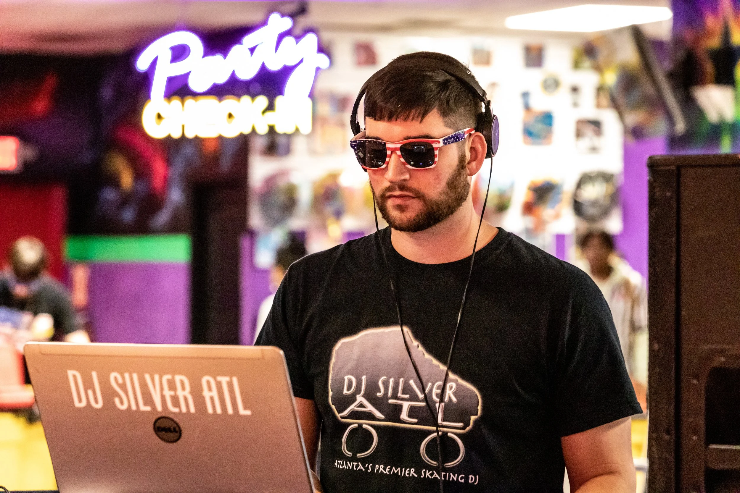 A man wearing sunglasses with an American flag design, headphones, and a black T-shirt with DJ Silver ATL written on it is DJing at an indoor skating rink with colorful lights and a neon sign in the background.