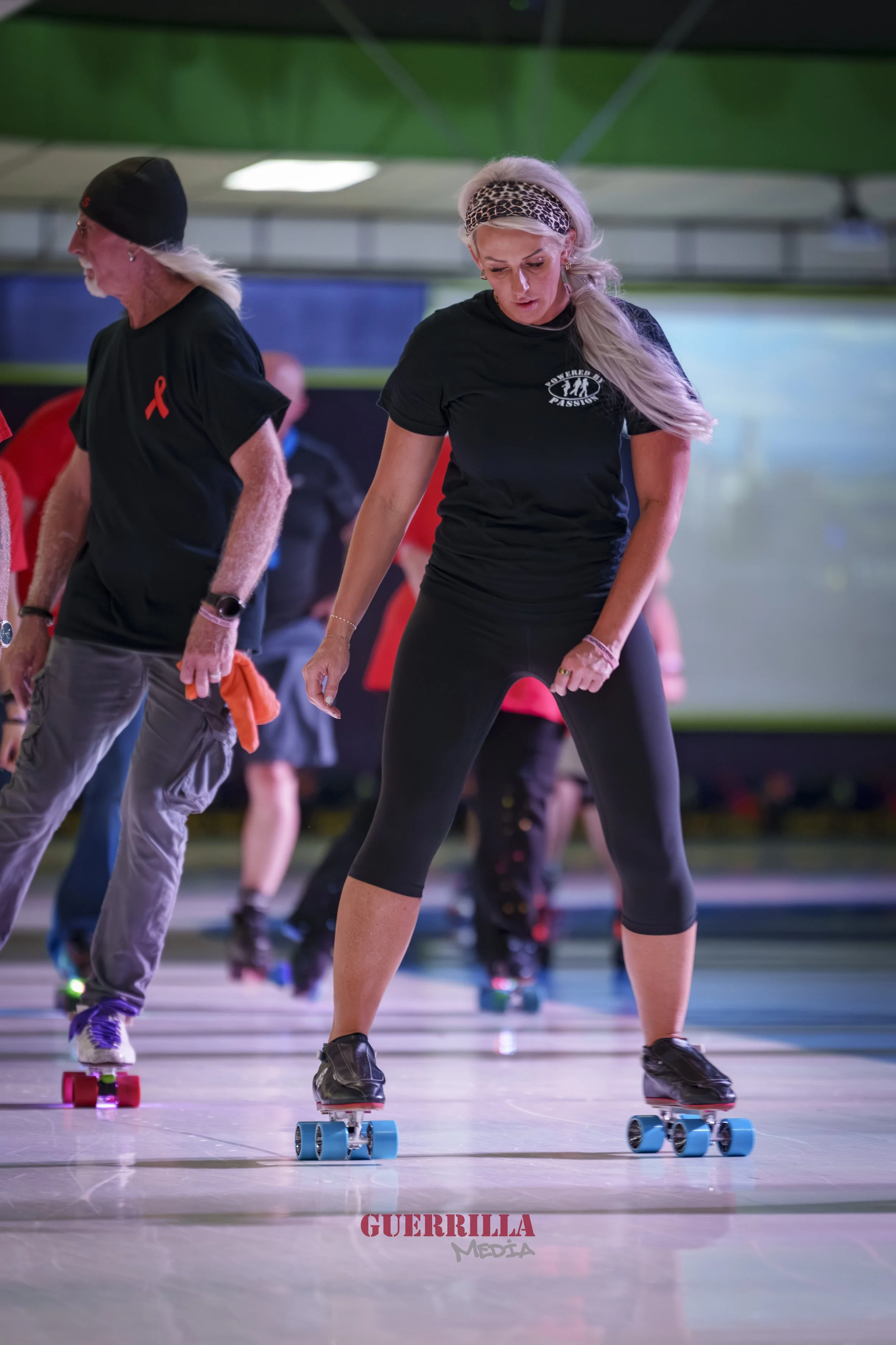 Group of women roller skating indoors, wearing black t-shirts and athletic clothing, with a focus on a blonde woman in the foreground looking down as she skates.