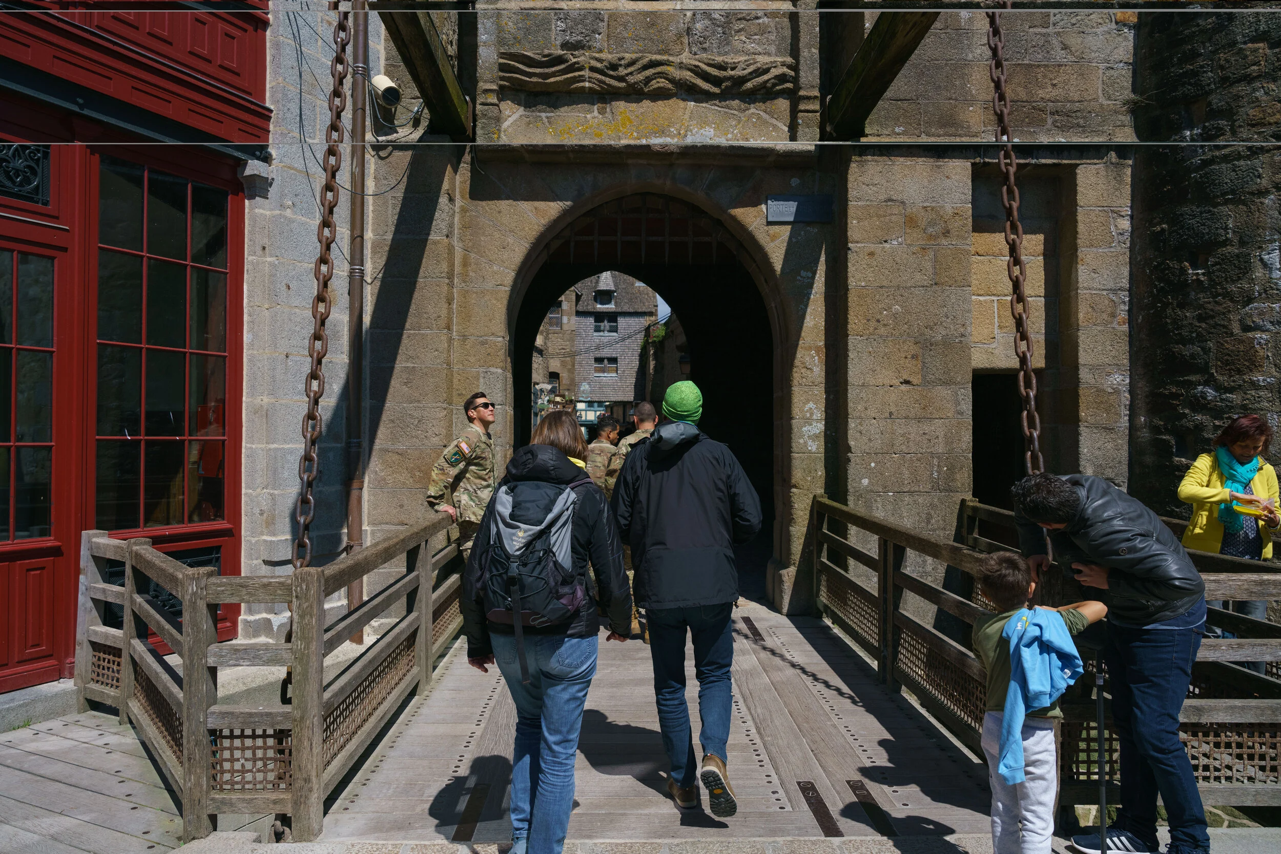 People walking through a stone archway in a historical site, with military personnel present and visitors taking photos on a sunny day.
