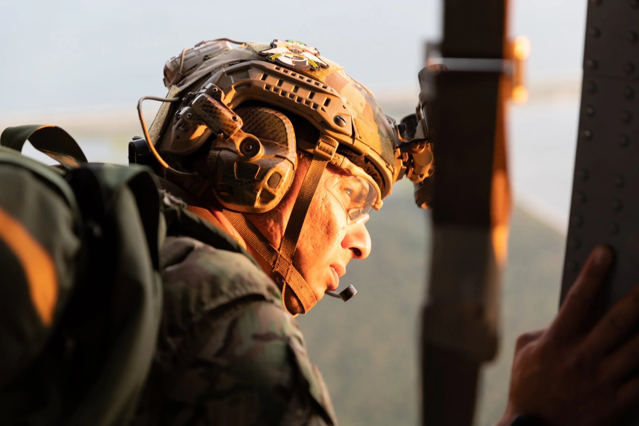 A soldier in camouflage uniform wearing a helmet with mounted communication gear, looking intensely at a touchscreen device.