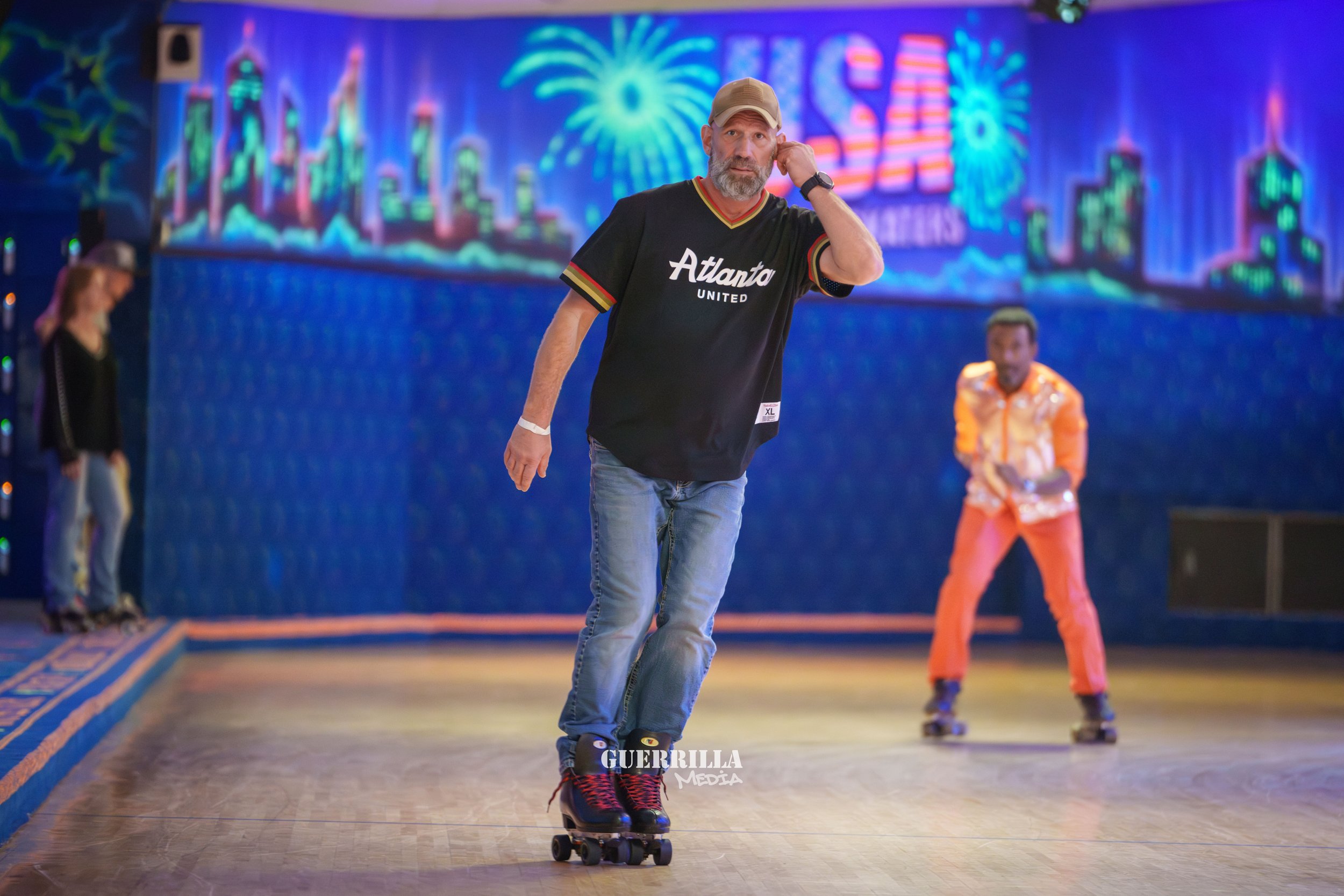 A man roller skating indoors with two women in the background, colorful neon cityscape and fireworks on a sign, and a vibrant blue wall behind. The man is wearing a black "Atlanta United" jersey, jeans, a cap, and roller skates.