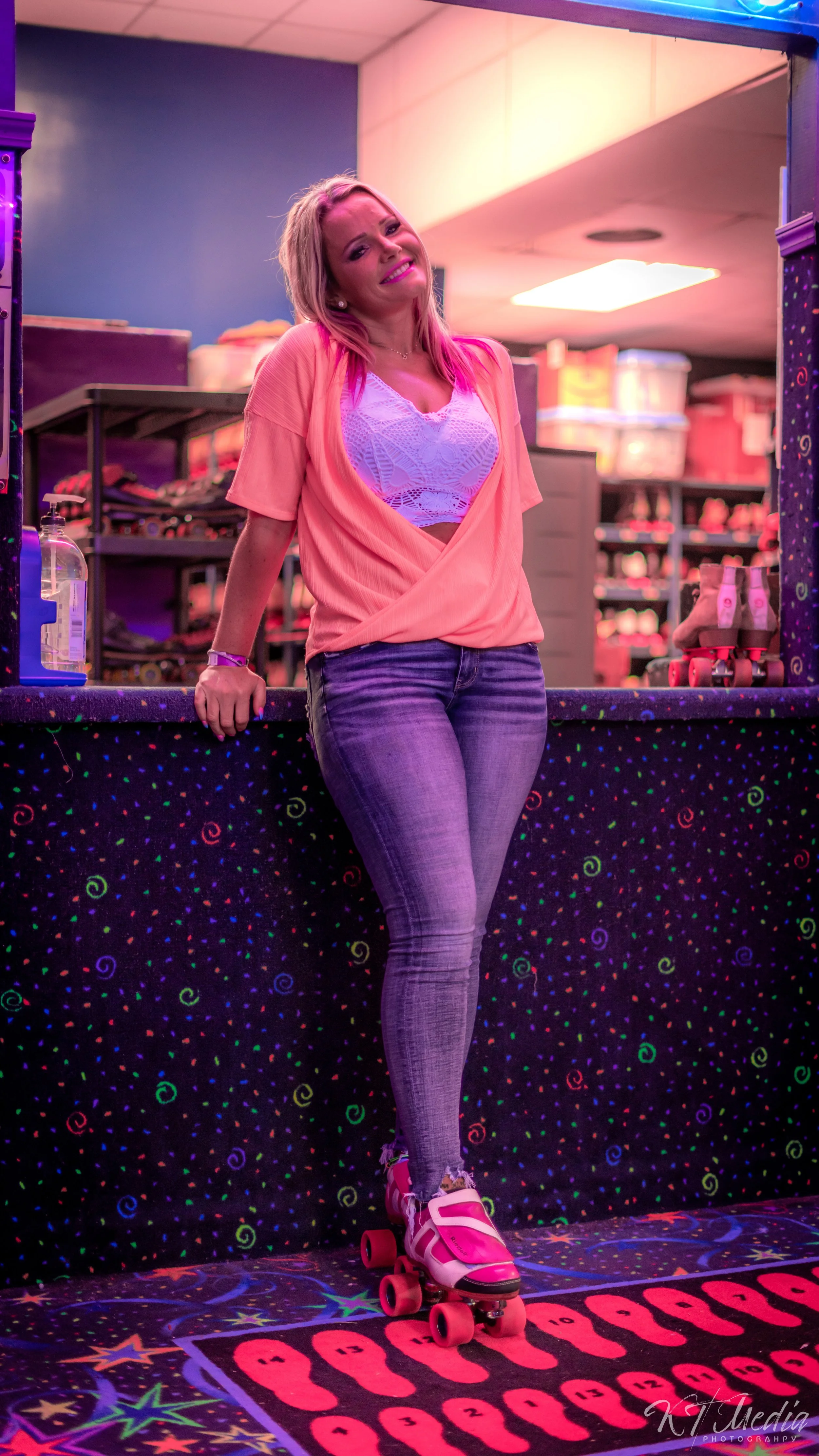 A woman in pink roller skates and casual clothing standing on a colorful, neon-lit roller skating rink floor with a wall and shelves of roller skates in the background.