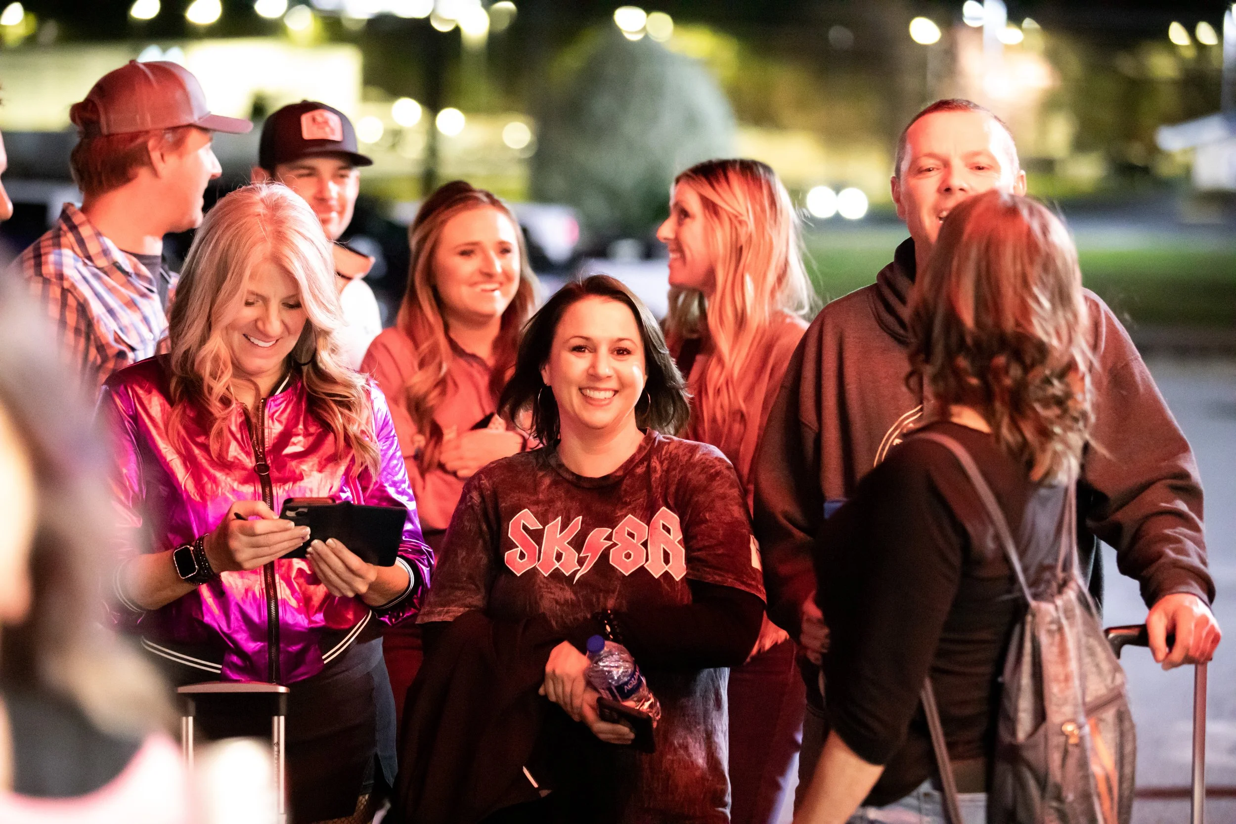 A group of people smiling and socializing outdoors during nighttime, with some holding water bottles and a suitcase, and one person checking their phone.