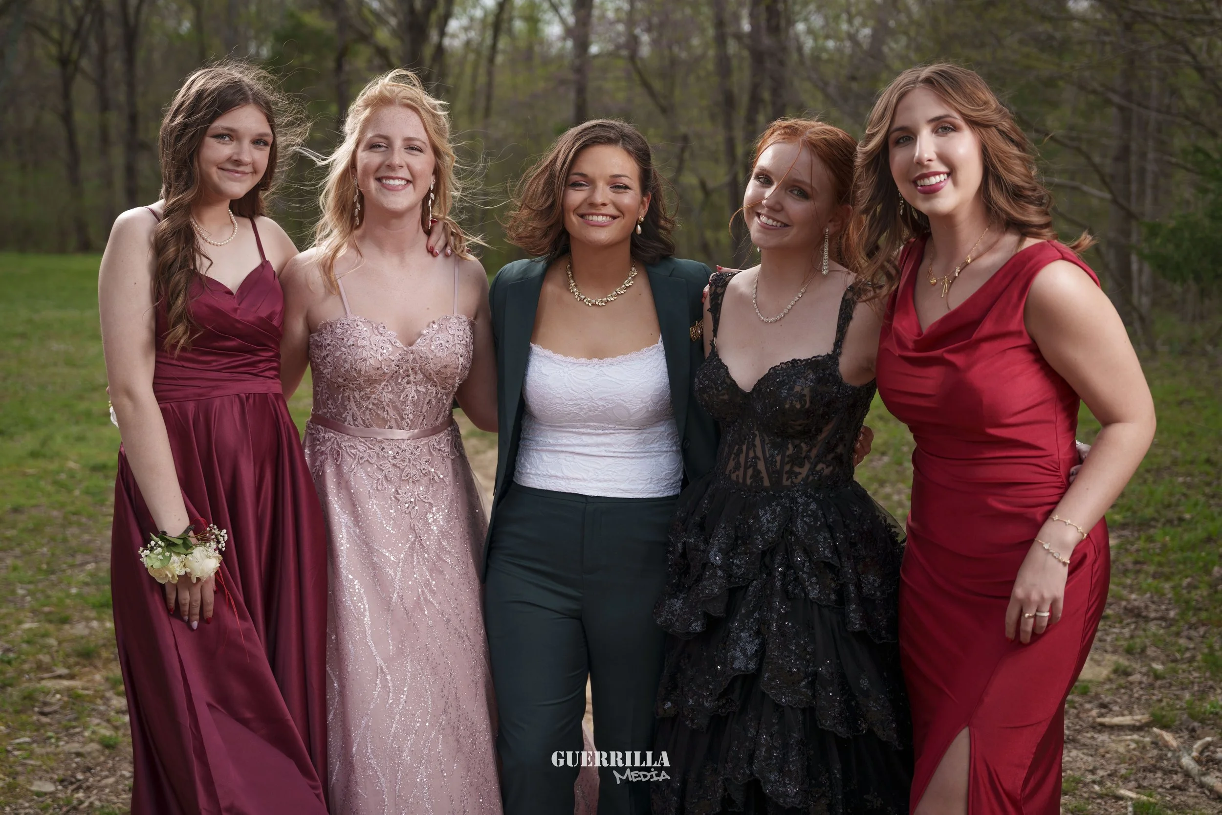 Group of five women dressed in formal attire standing outdoors in a wooded area, smiling for the camera.