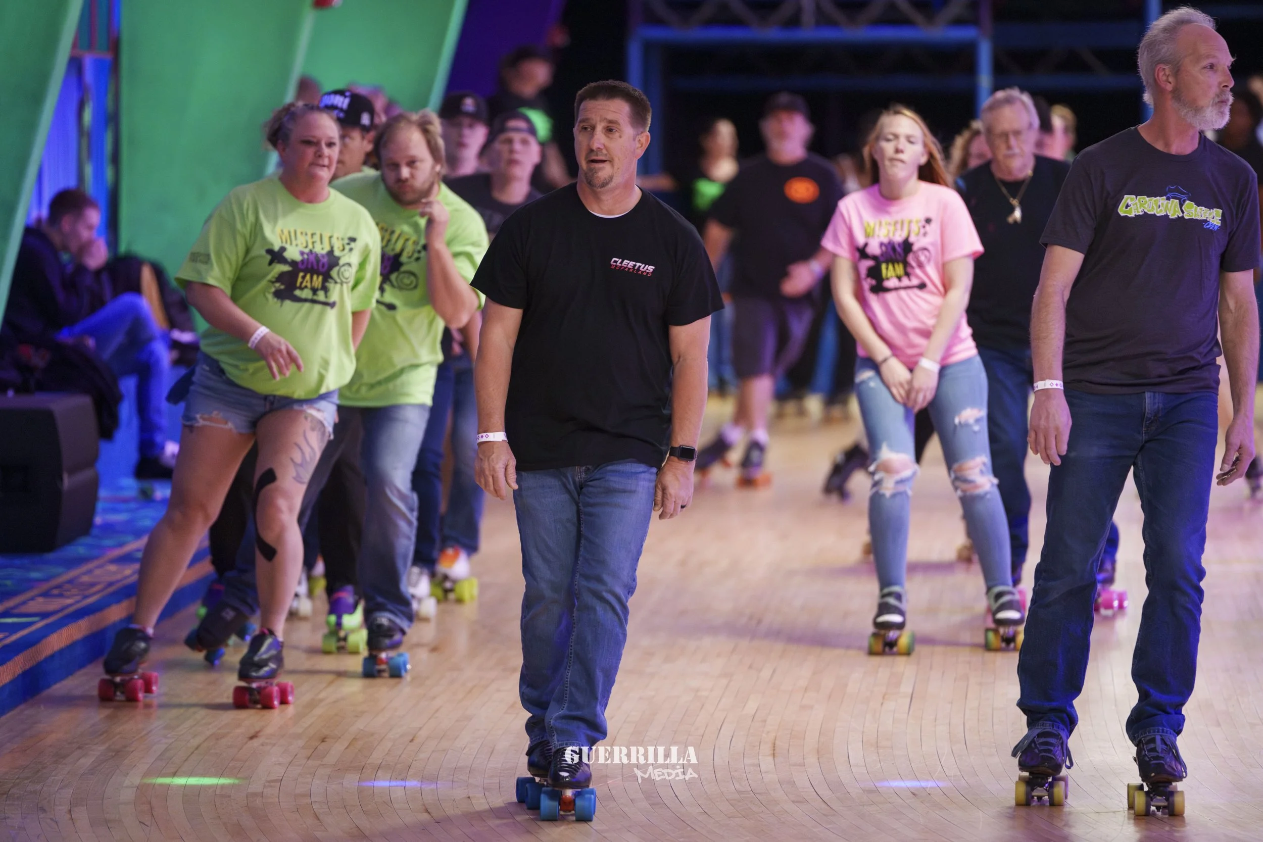 People roller skating indoors at a recreational event, with some wearing colorful t-shirts, in a large skating rink.