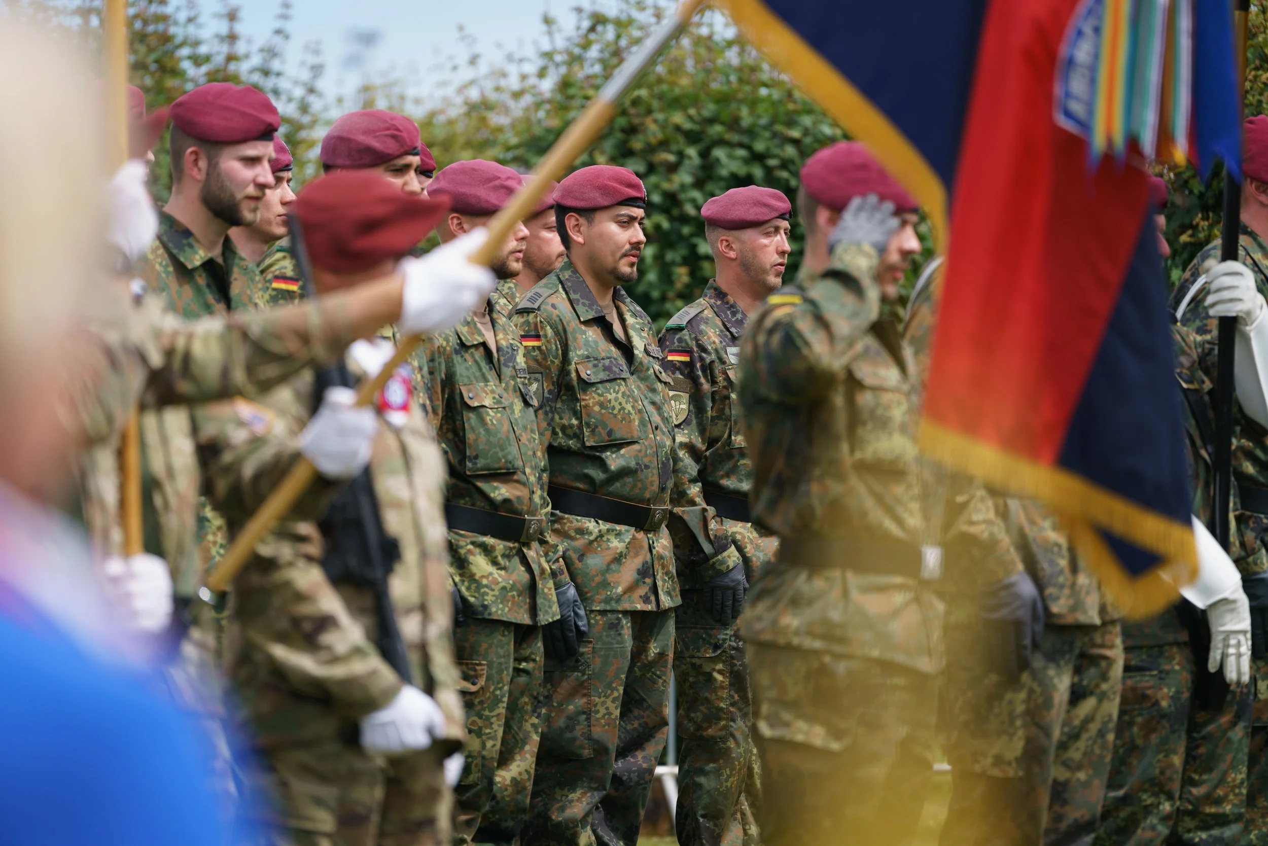 Group of soldiers in camouflage uniforms and maroon berets standing in formation during a ceremony, with some holding flags.