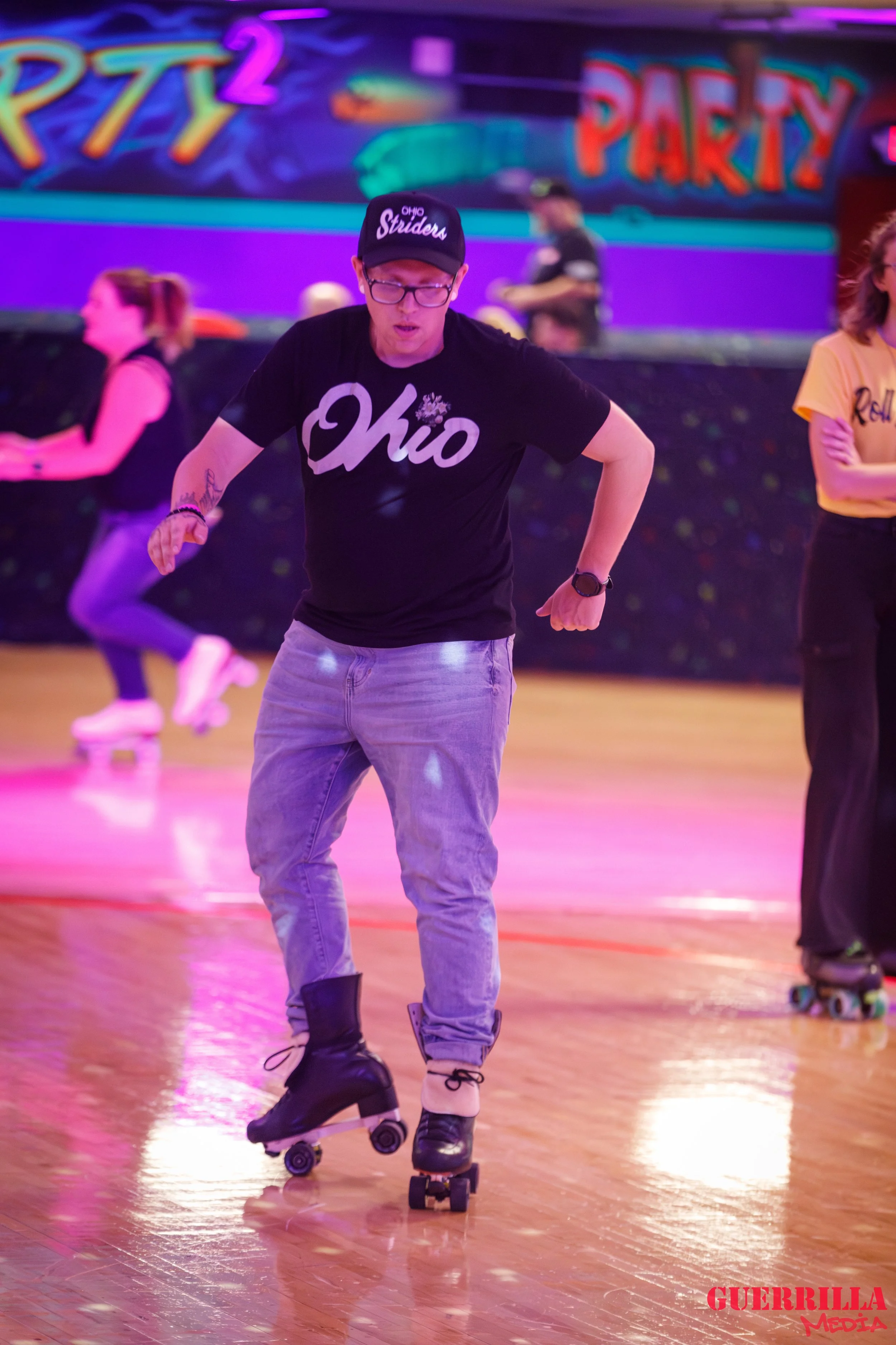 A person roller skating indoors under colorful neon lights, wearing a black 'Ohio' T-shirt, light-colored jeans, and a black cap with white text, at an indoor roller rink with a vibrant background that says 'PARTY' and 'RPT2'.