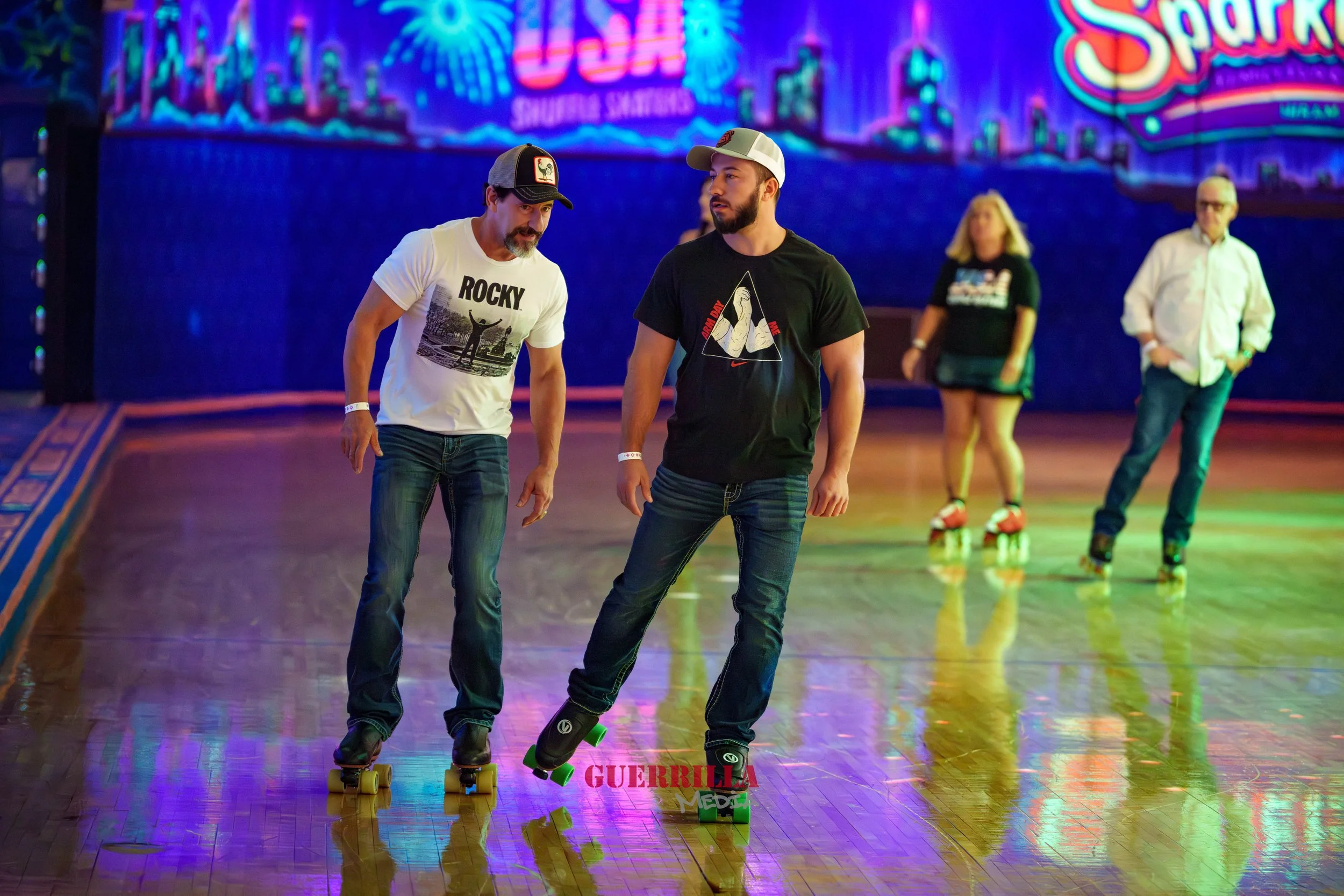 Two men roller skating in an indoor rink, dressed casually with hats, one wearing a "Rocky" t-shirt and the other a black t-shirt with a graphic. In the background, two other people are skating, a woman in a black shirt and shorts, and a man in a whi