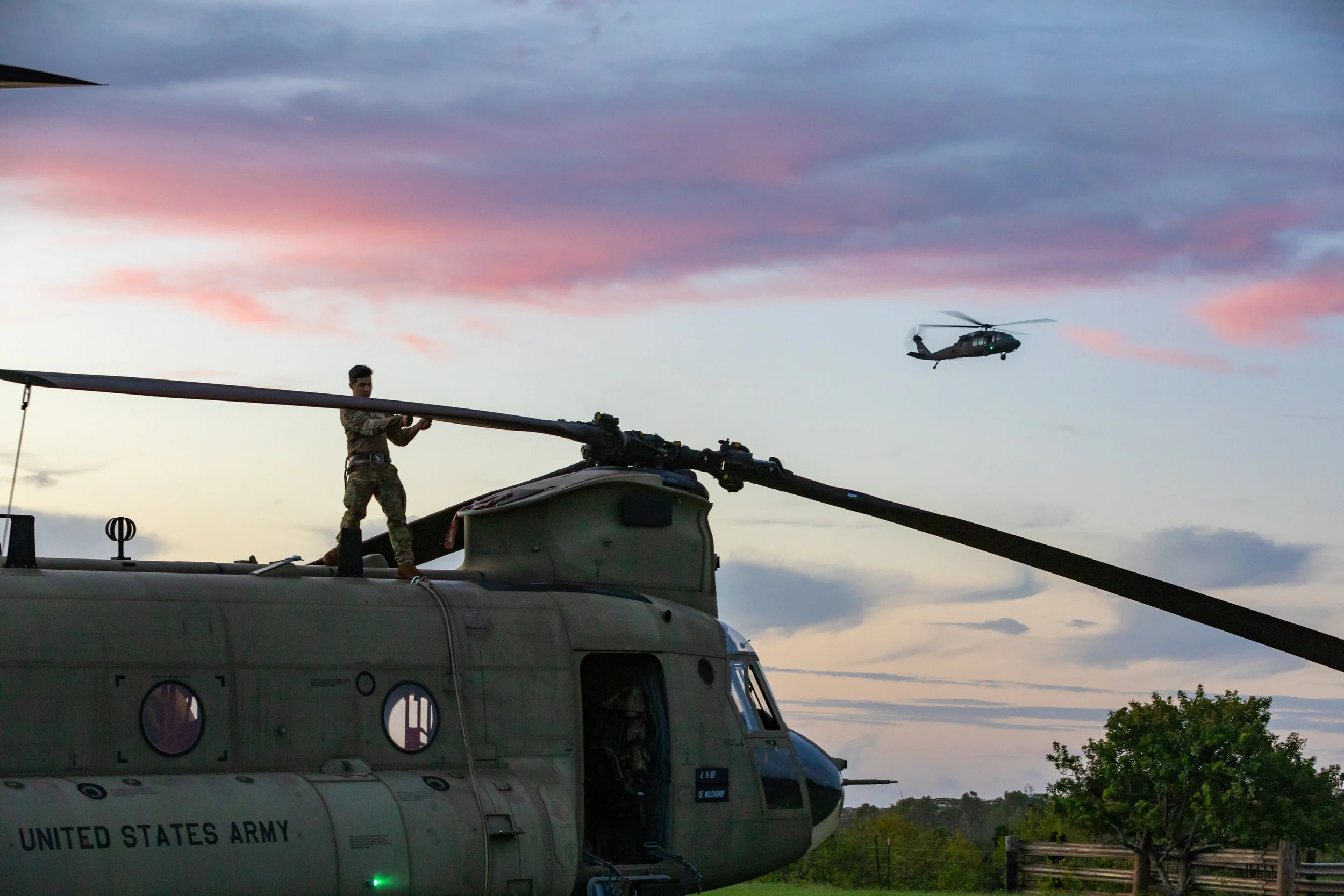 A United States Army helicopter with a soldier standing on top, preparing the rotor blade, during sunset. A second helicopter is flying in the sky in the background.
