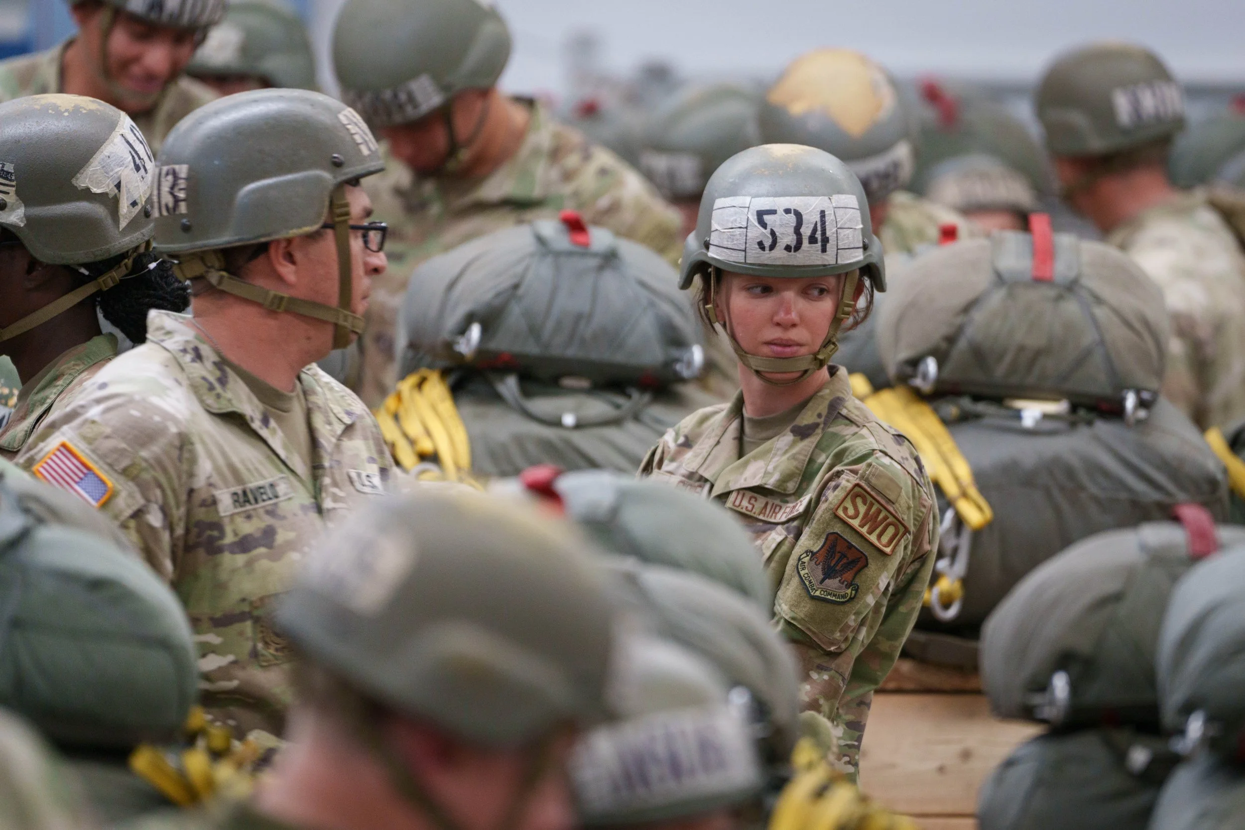 A group of soldiers in uniform and helmets standing together, with some backpacks visible in the background.