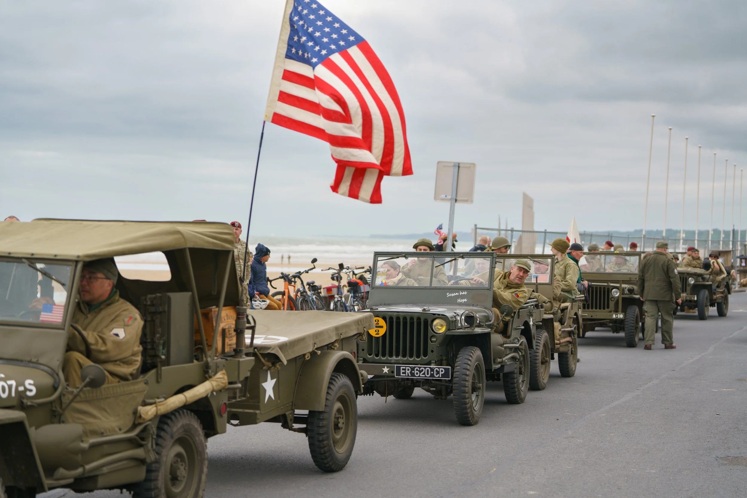 Vintage military vehicles decorated with American flags parked along a beach promenade with soldiers and civilians nearby.