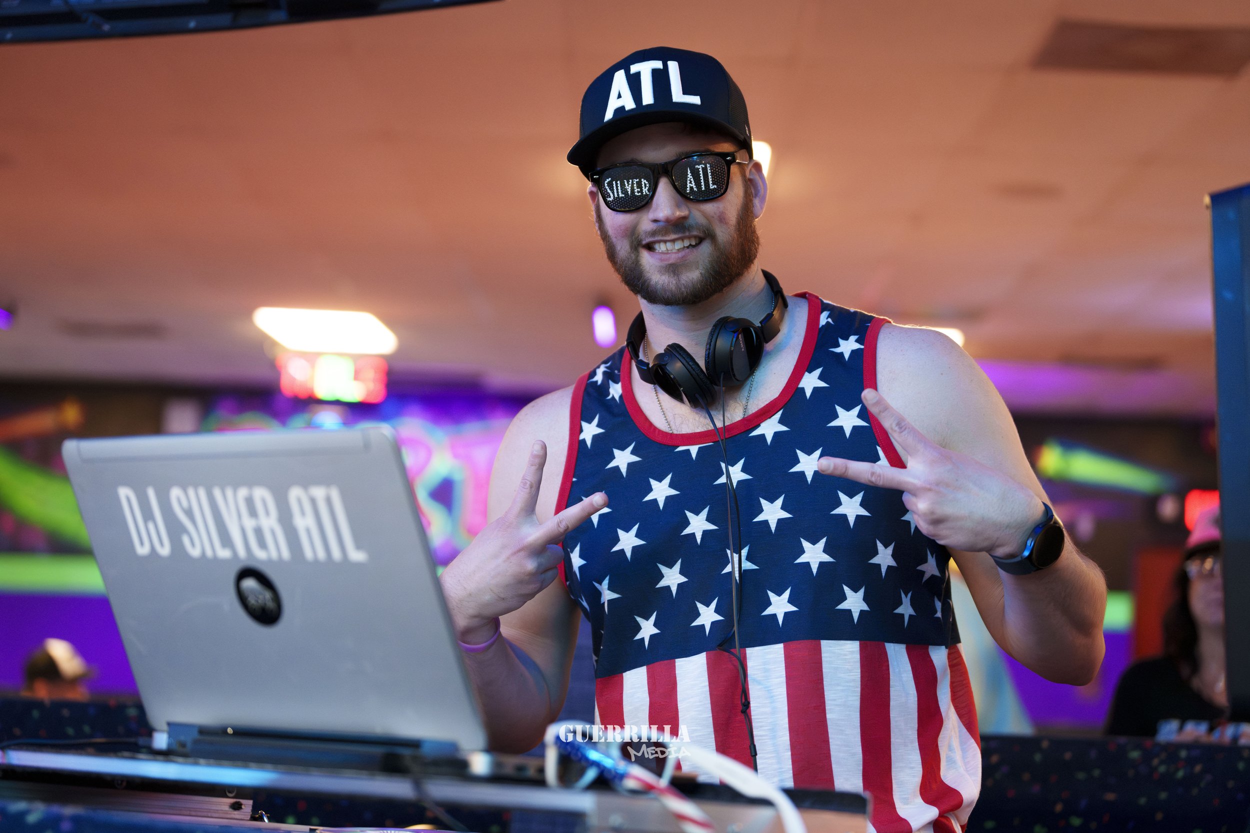 DJ wearing a black cap with white 'ATL' letters, sunglasses with 'SILVER' and 'ATL', and a patriotic tank top with stars and stripes, smiling and making peace signs at a colorful party.