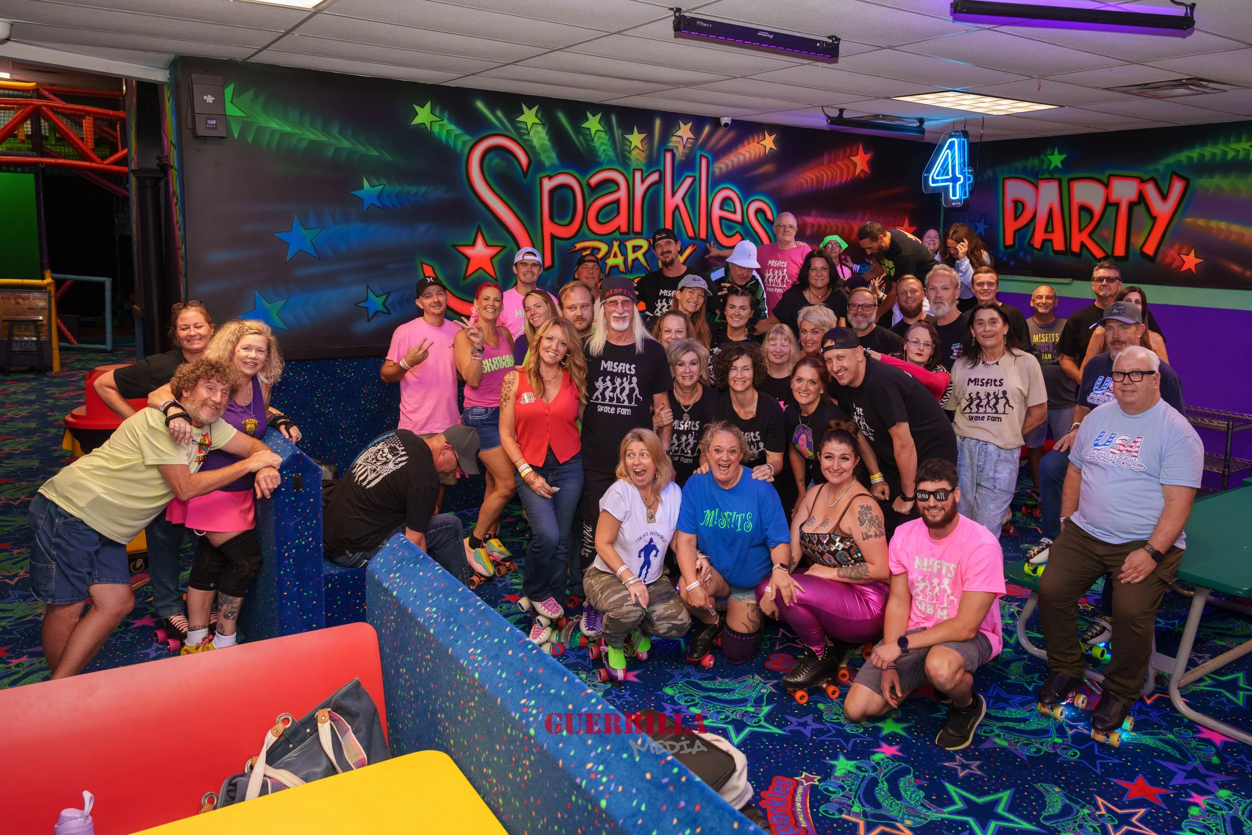A group of people posing for a photo at a roller skating rink with a colorful, neon-lit backdrop that reads "Sparkle Party." Some are wearing skate gear and colorful outfits, smiling and having fun.