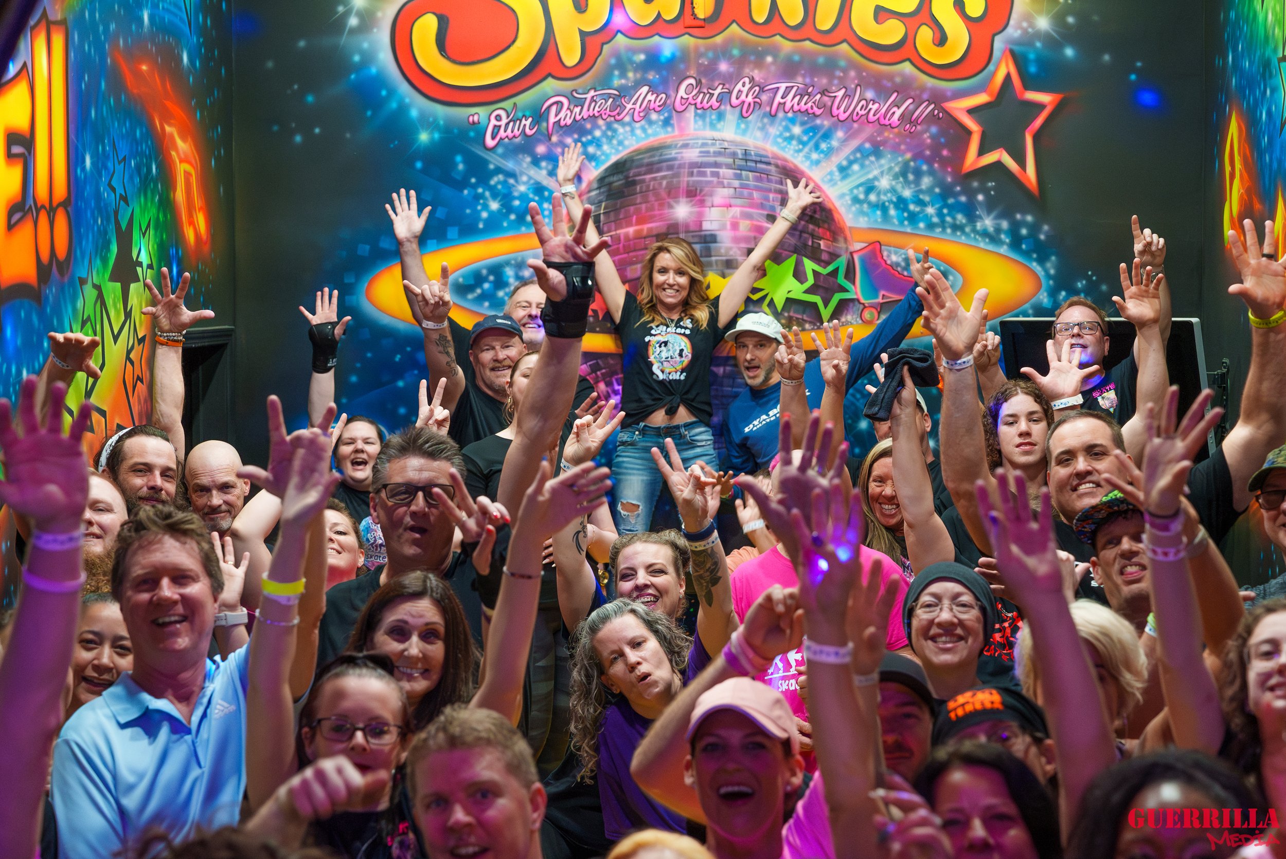 A large group of people at a lively event, with many raising their hands and smiling. In the background, a colorful mural features the word 'Space' and a slogan 'Our Parties Are Out Of This World!!' along with stars and a disco ball. The crowd appear