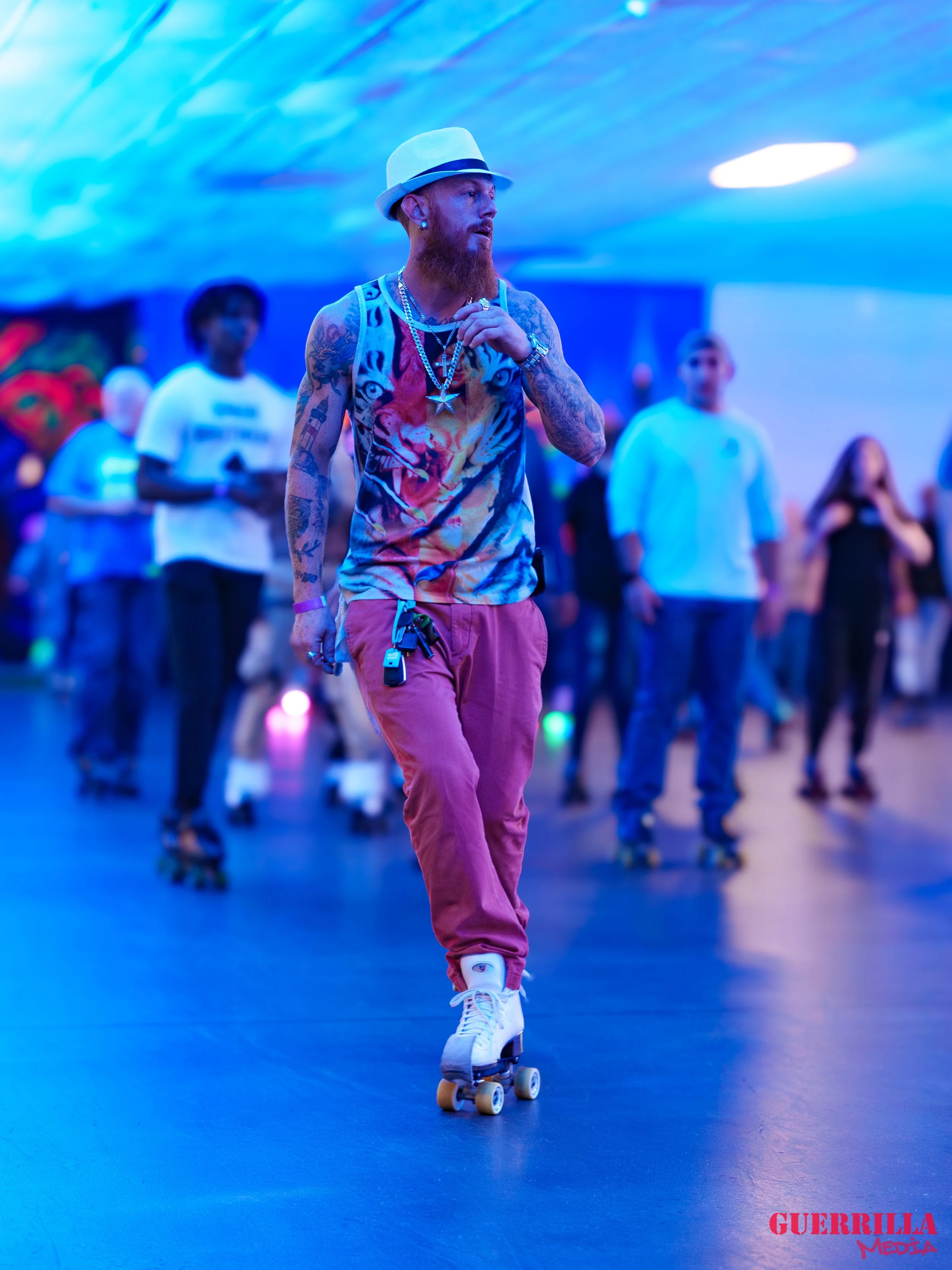 A man with tattoos and a red beard roller skating in an indoor skate rink with colorful lighting, wearing a hat, a tiger print tank top, pink pants, and jewelry.