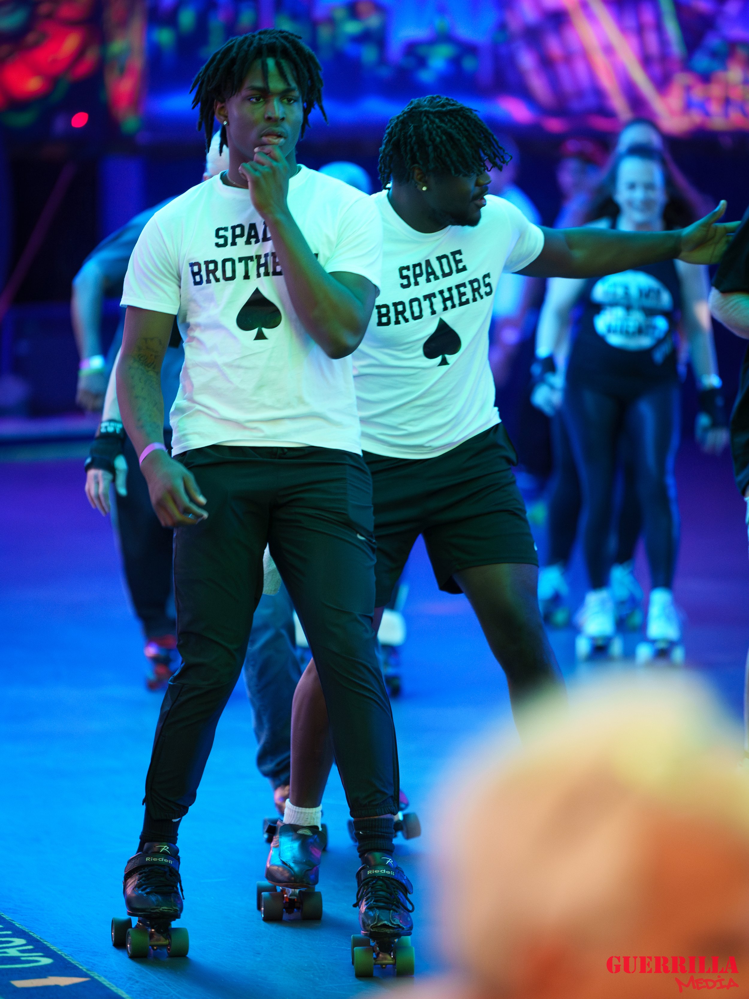 Two young men roller skating at an indoor skate rink with colorful neon lighting. They are wearing matching white t-shirts that read 'Spade Brothers' with a black spade symbol. One has his hand on his chin, and the other is extending his arm.