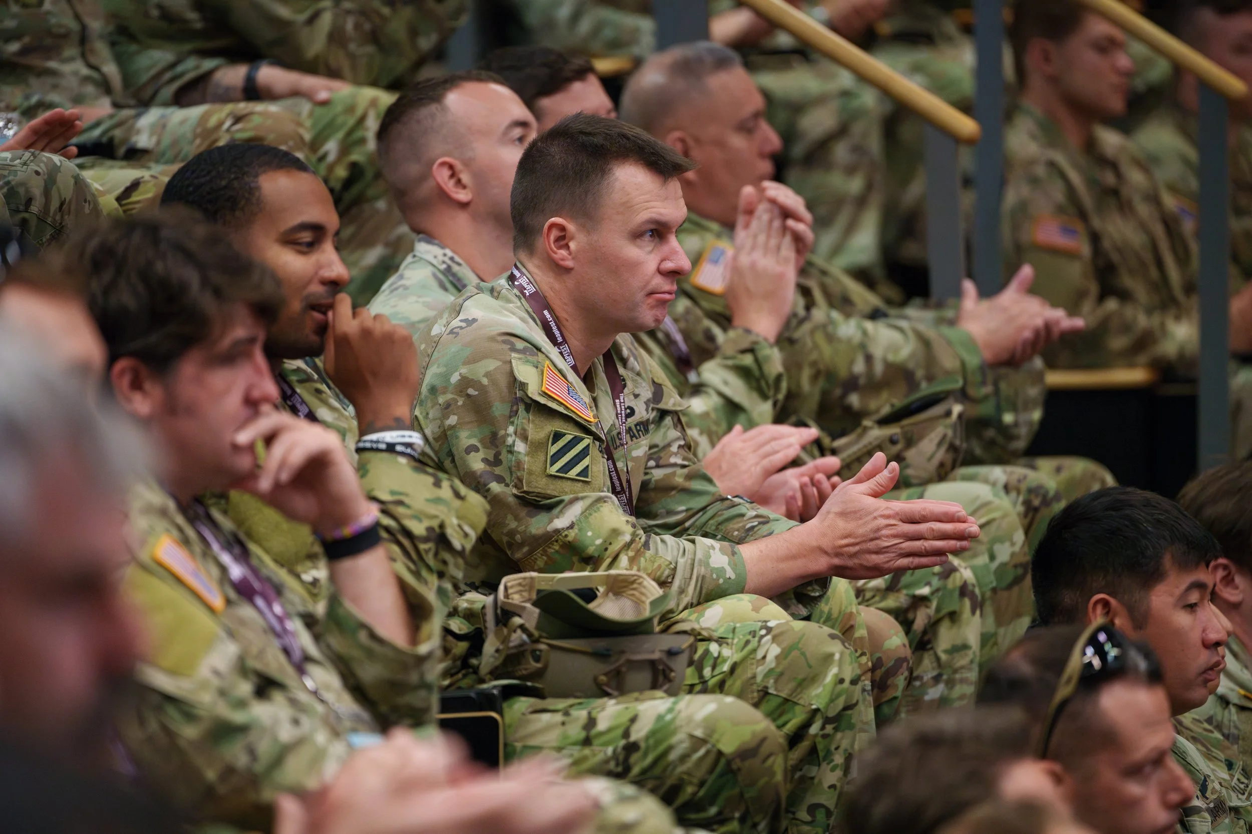 A group of soldiers in camouflage uniforms sitting in an indoor auditorium, attentively listening or watching a presentation, with some clapping and others with hands clasped.