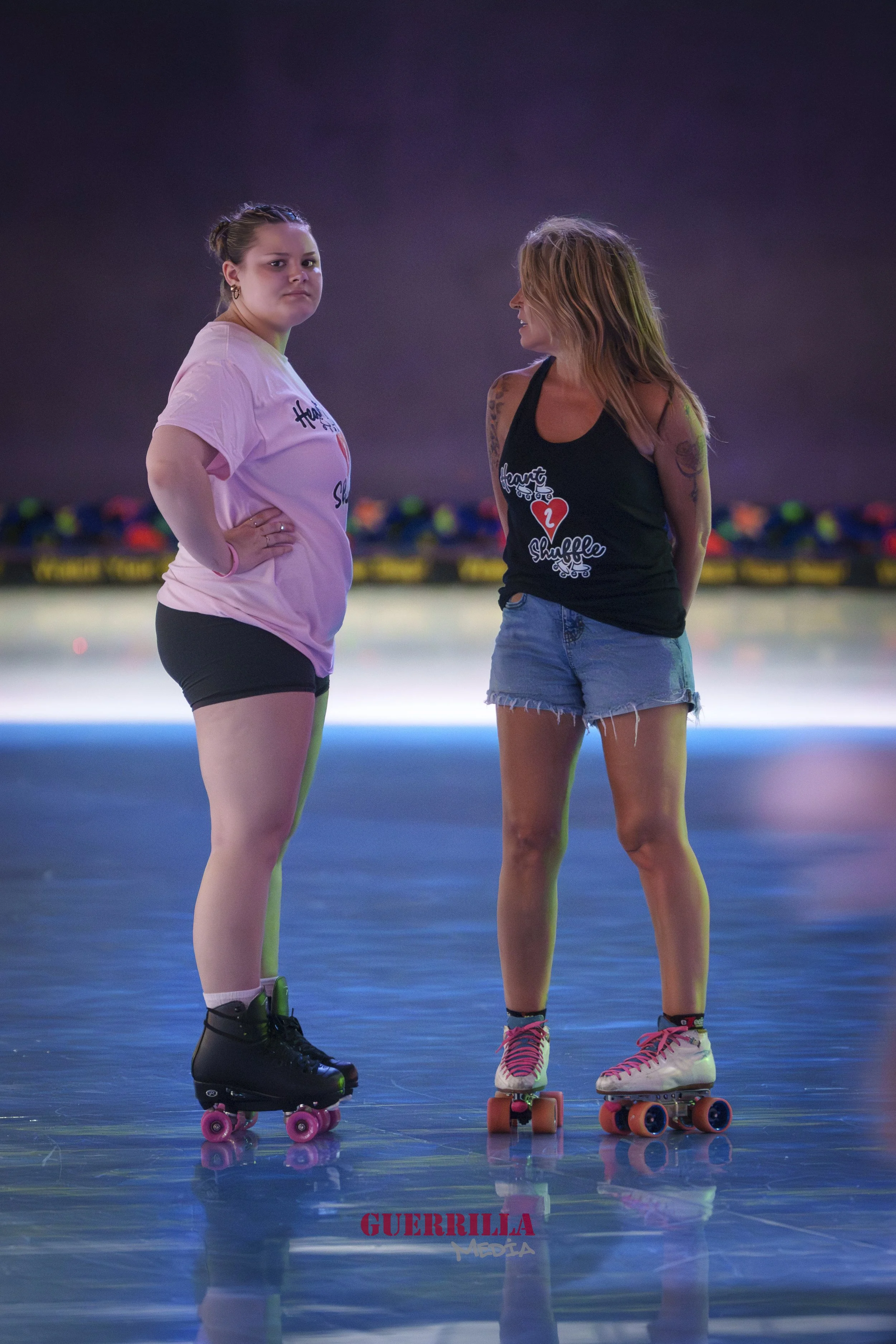 Two women wearing roller skates standing in a roller skating rink, facing each other with one woman looking serious and the other smiling.