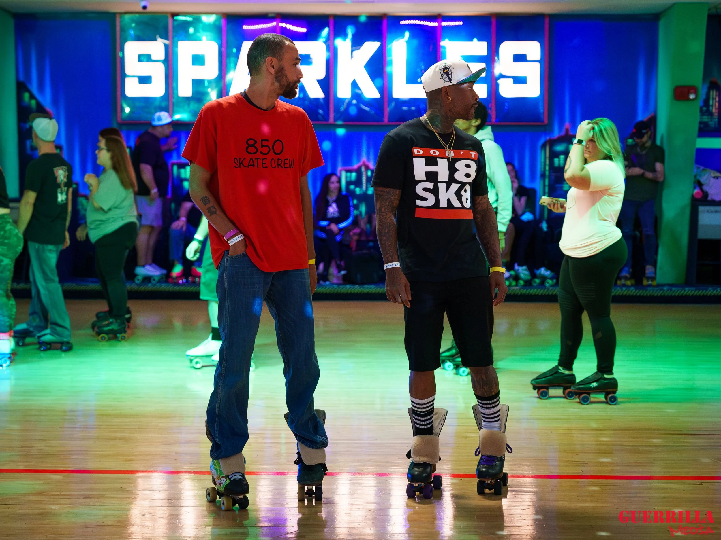 Two men roller skating in an indoor rink with other skaters and a neon sign that says 'Sparkles' in the background.