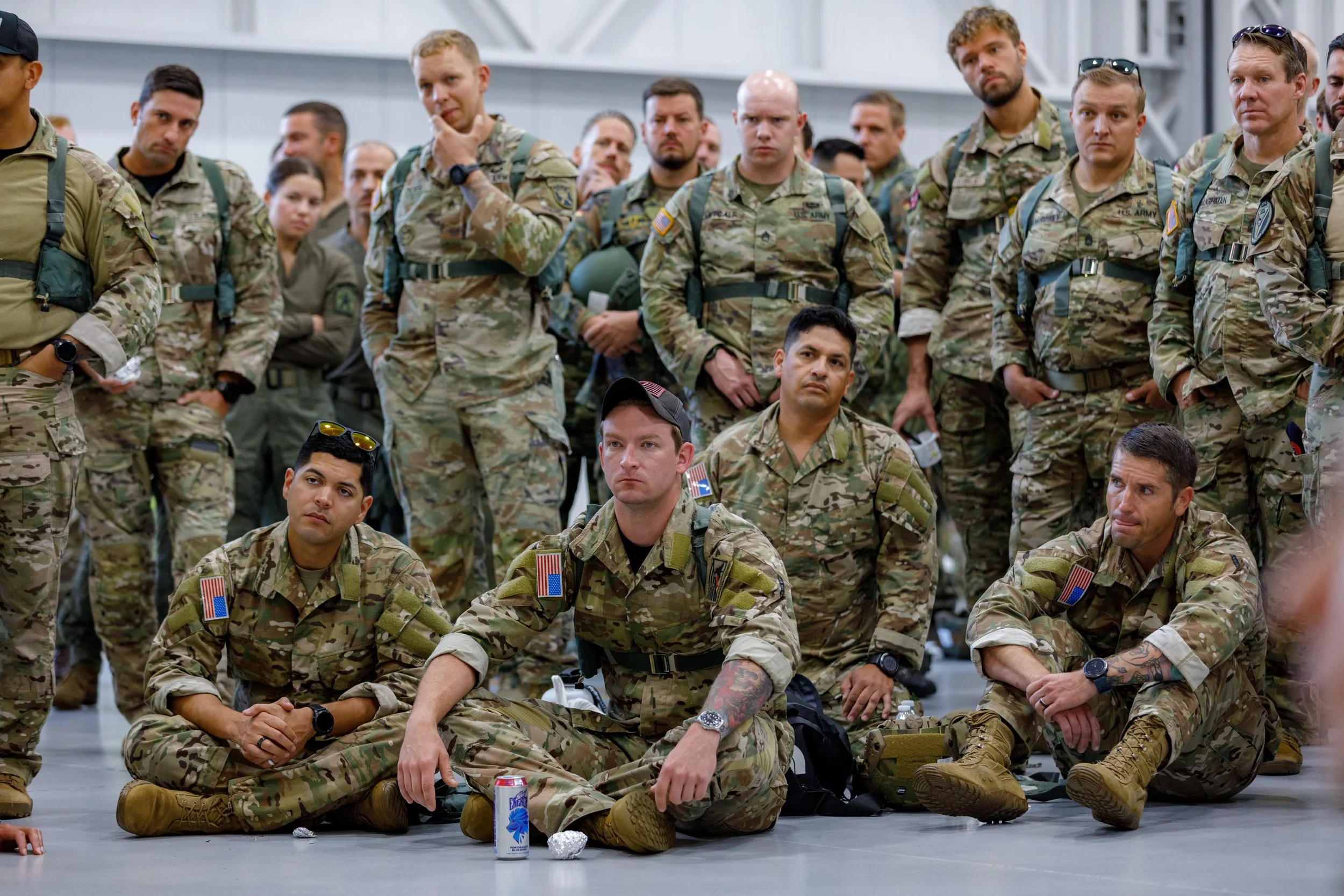 Group of soldiers in camouflage uniforms, some sitting on the floor and others standing, inside a large indoor space.