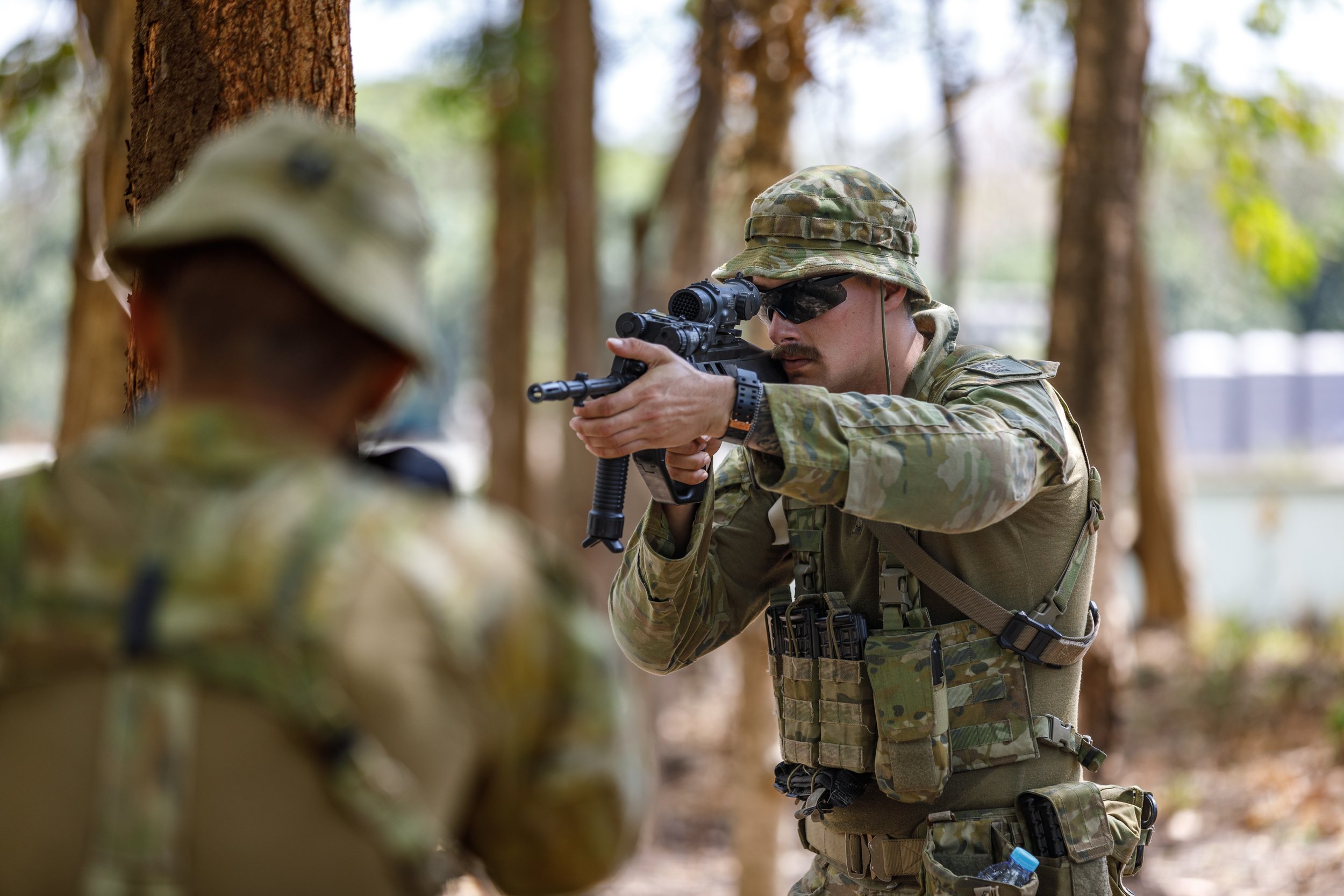 A soldier aiming a rifle in a wooded area, wearing camouflage gear, sunglasses, and a helmet.