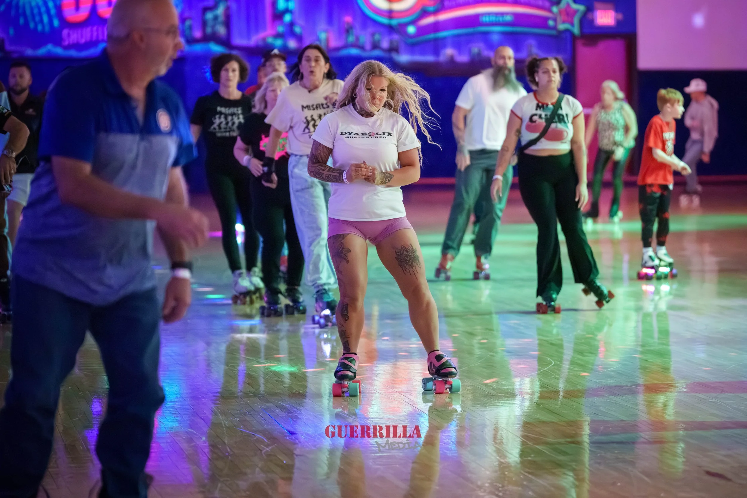 People roller skating at an indoor rink with colorful neon lights, including a woman with long blonde hair, tattoos, and a white t-shirt, in the foreground.