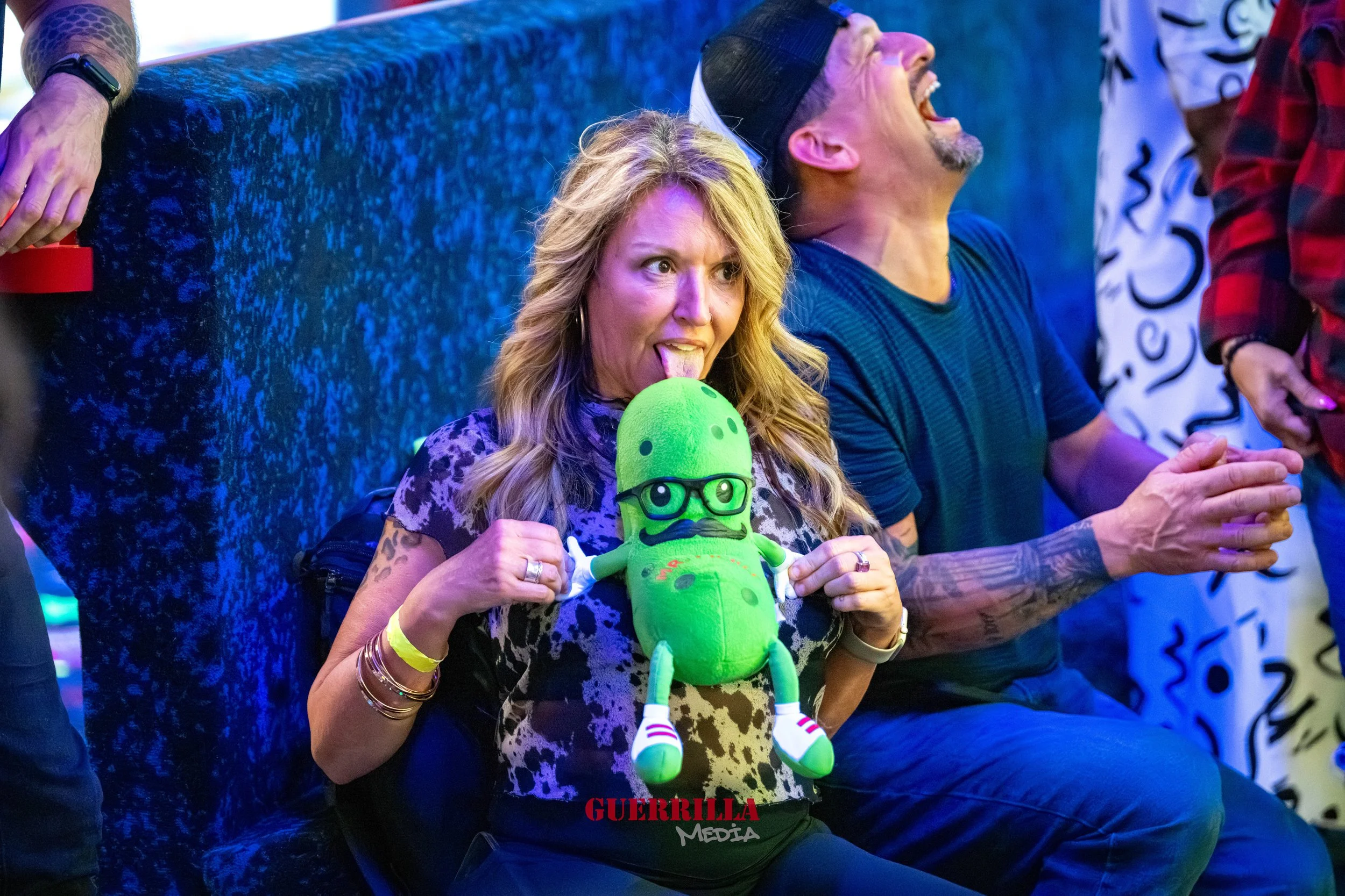 Woman with wavy blonde hair holding a green plush toy with glasses and a mustache, licking the toy's face, while a man beside her claps and laughs at a party or event.