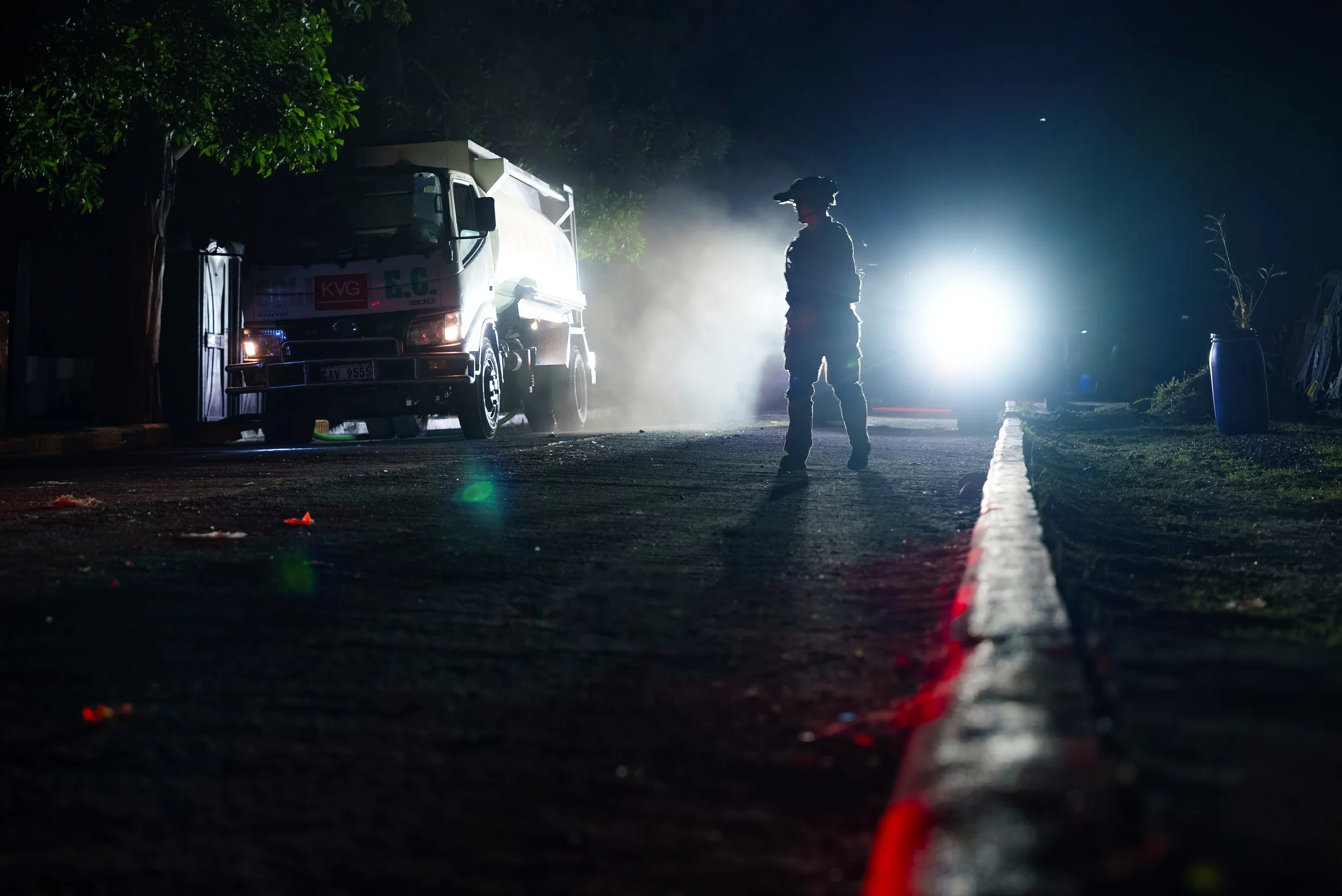 A Soldier walking on a dark street at night with a bright light behind him, a truck nearby, and smoke in the air.